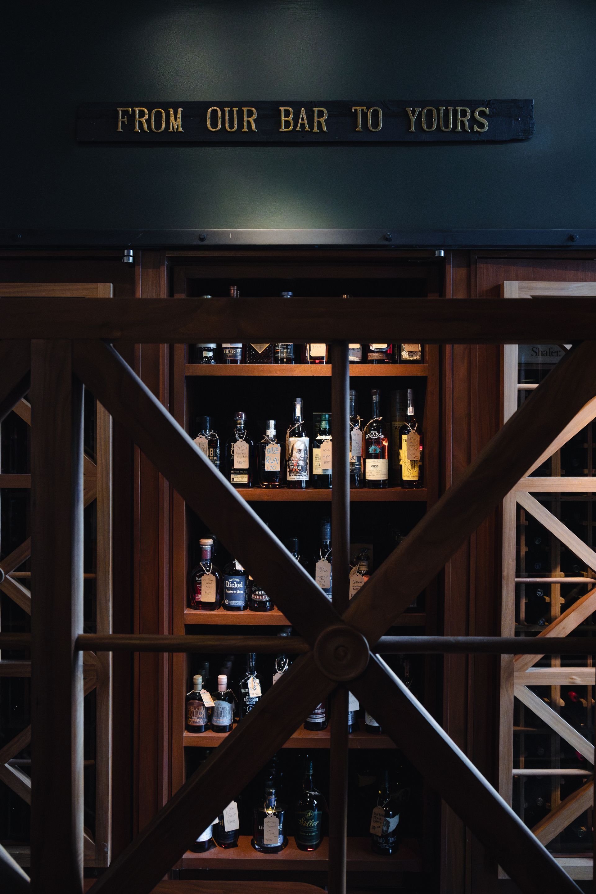 Dim bar shelf with liquor bottles behind wooden cross-brace doors and a sign reading “From Our Bar to Yours”