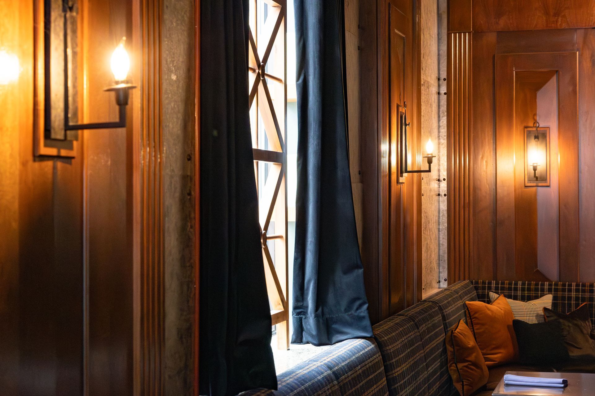 Cozy room with wood paneling, sconce lighting, and a window with dark curtains. A booth with pillows is visible.