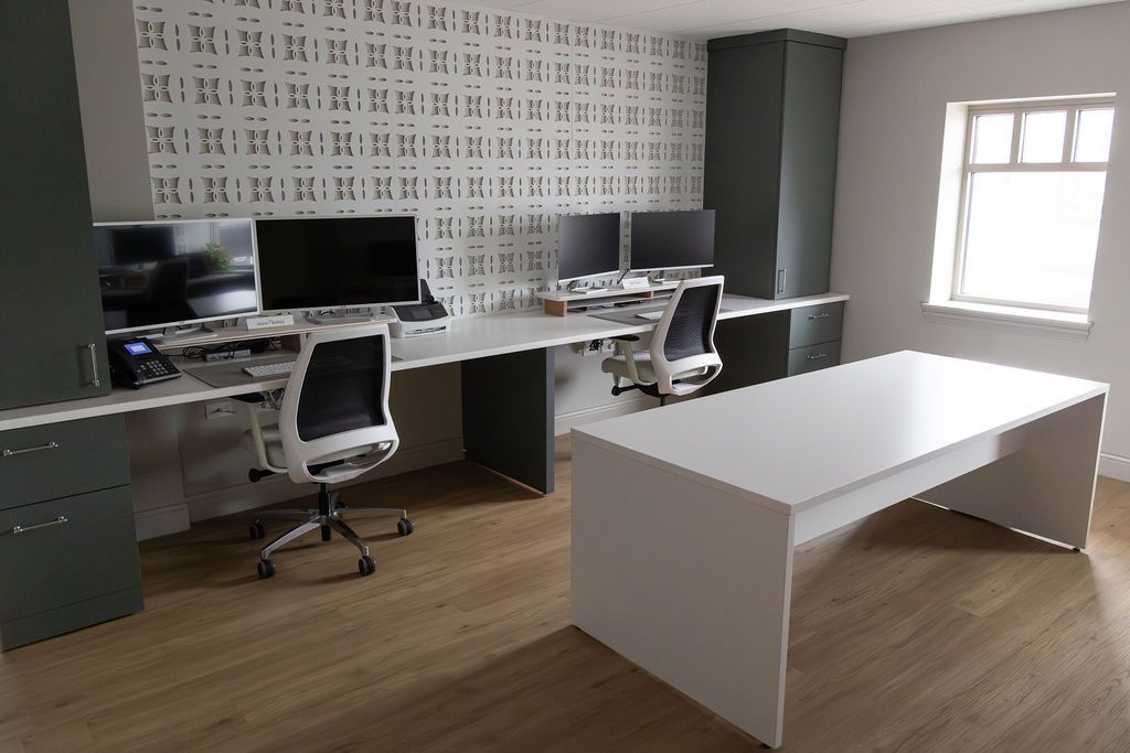 Modern office with multiple computer desks and chairs, white walls, and a large window.