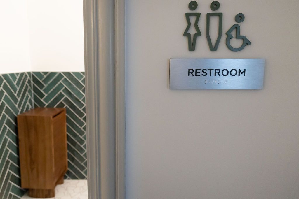 Restroom sign with gender symbols and accessibility icon on a light gray wall, doorway to the left.