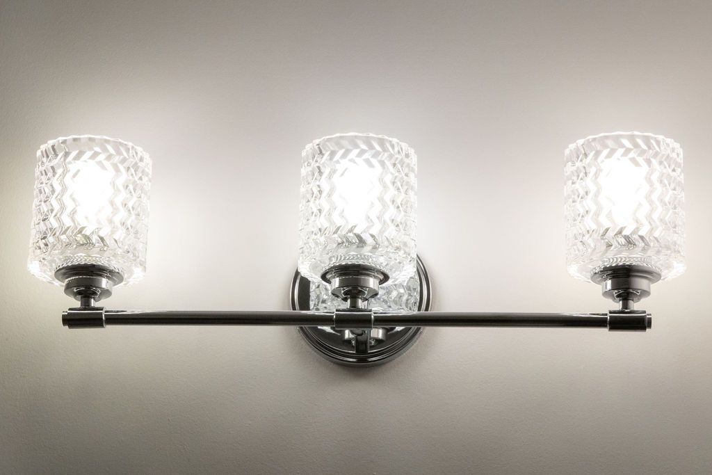 Three-light bathroom vanity fixture with textured glass shades and dark metal arms, mounted on a light wall.