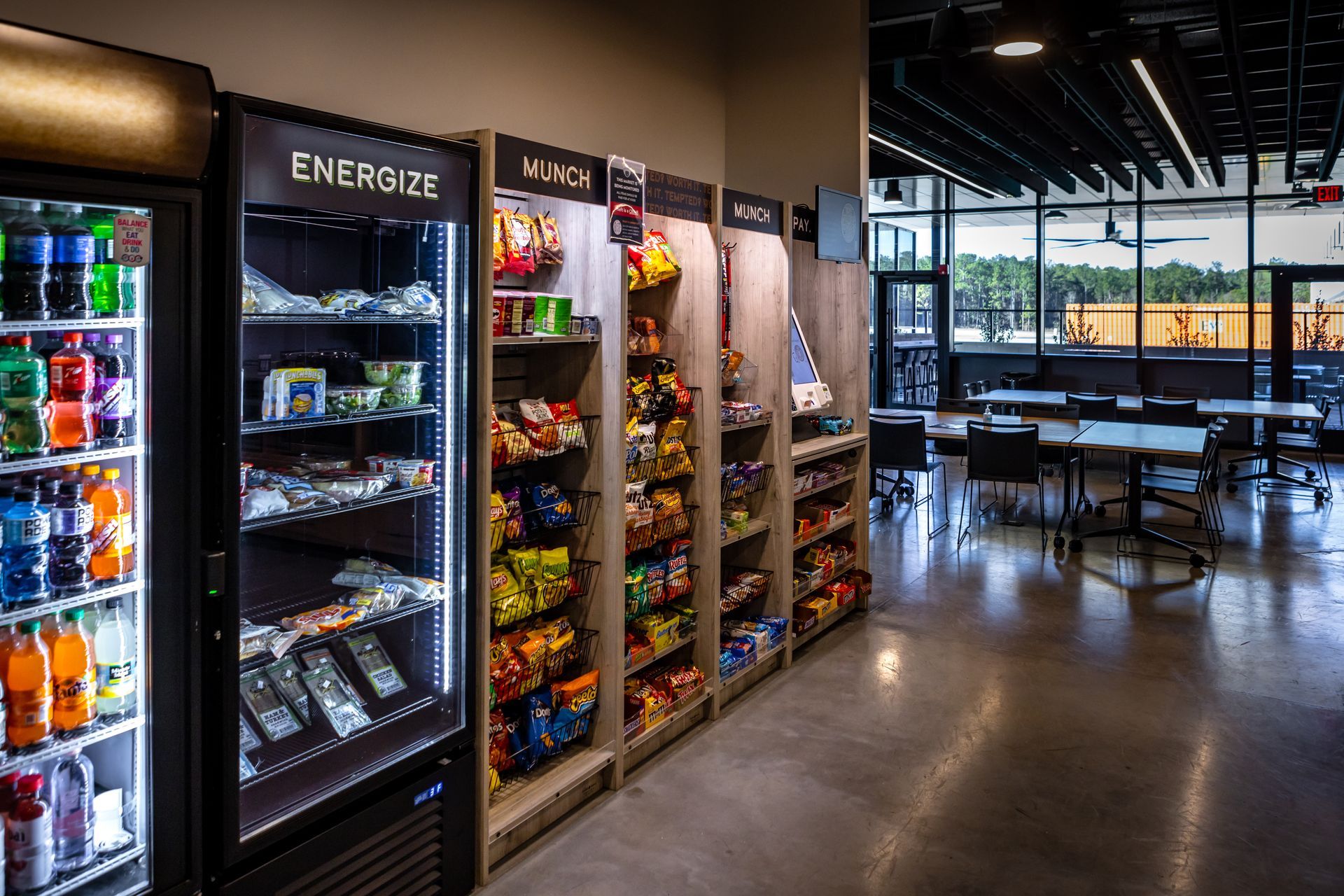A vending area with a drink cooler, snack shelves, and tables in a brightly lit space.