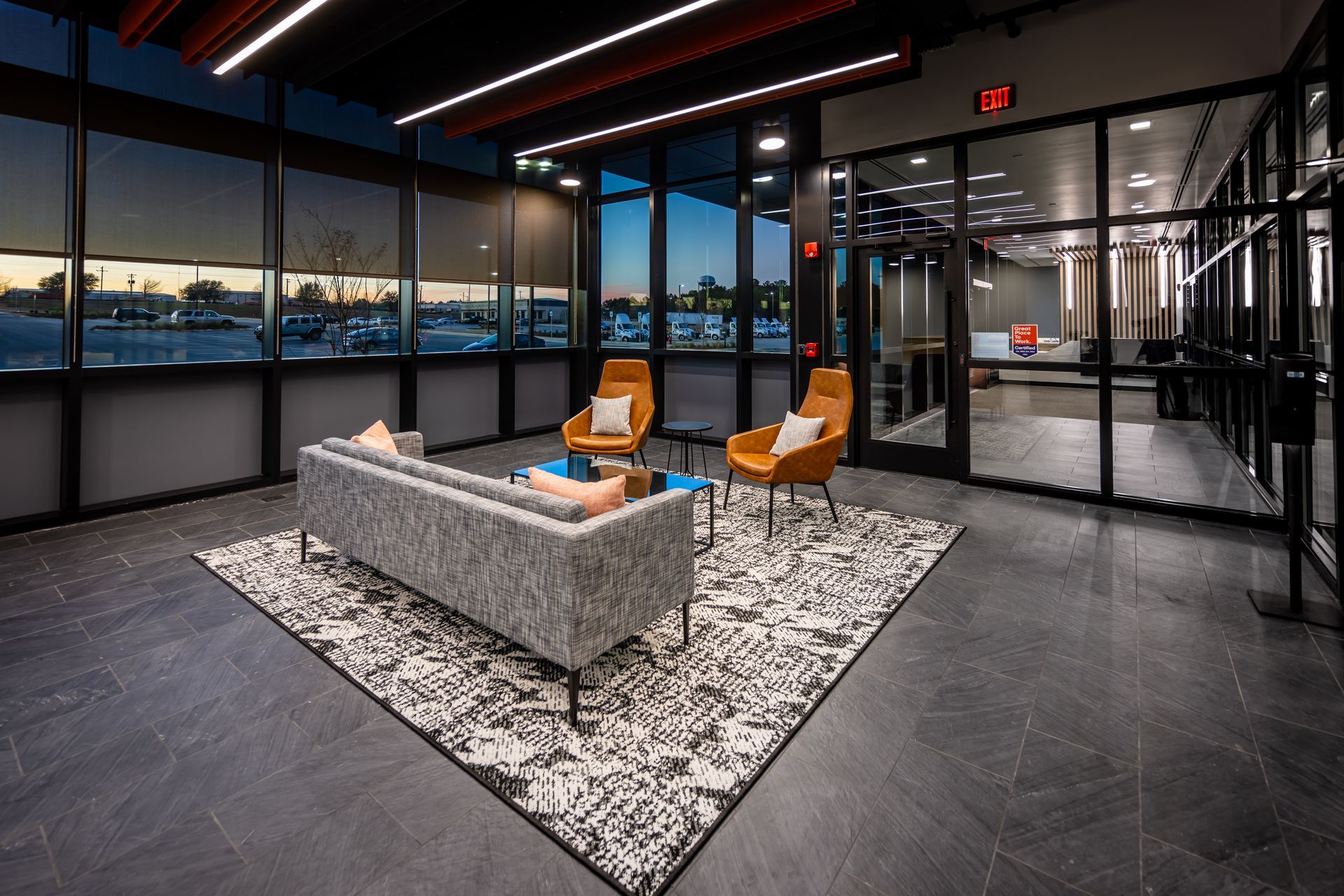 Modern office waiting area with dark gray flooring, patterned rug, sofa, two chairs, and large windows.