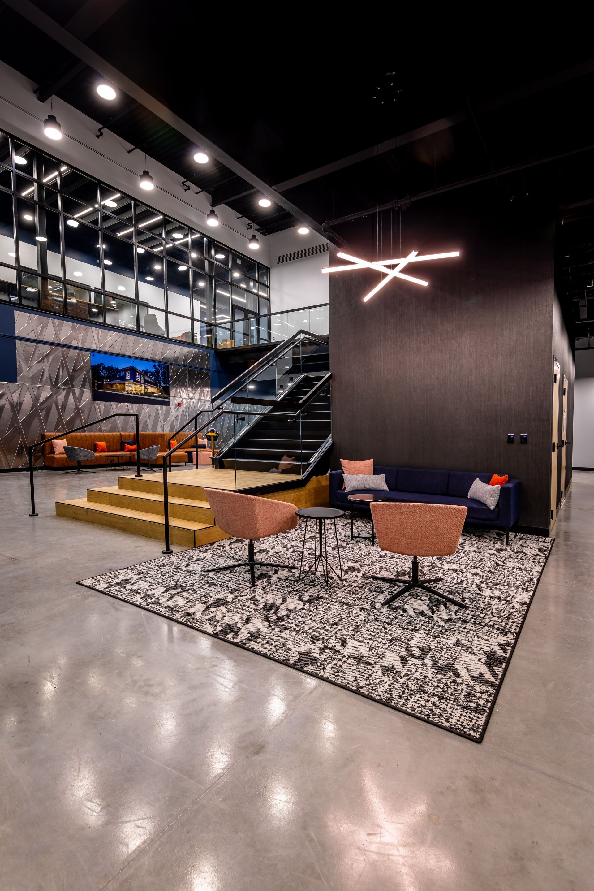 Modern office lounge with stairs, seating area, and artwork. Concrete floor, black ceiling, and accent lighting.