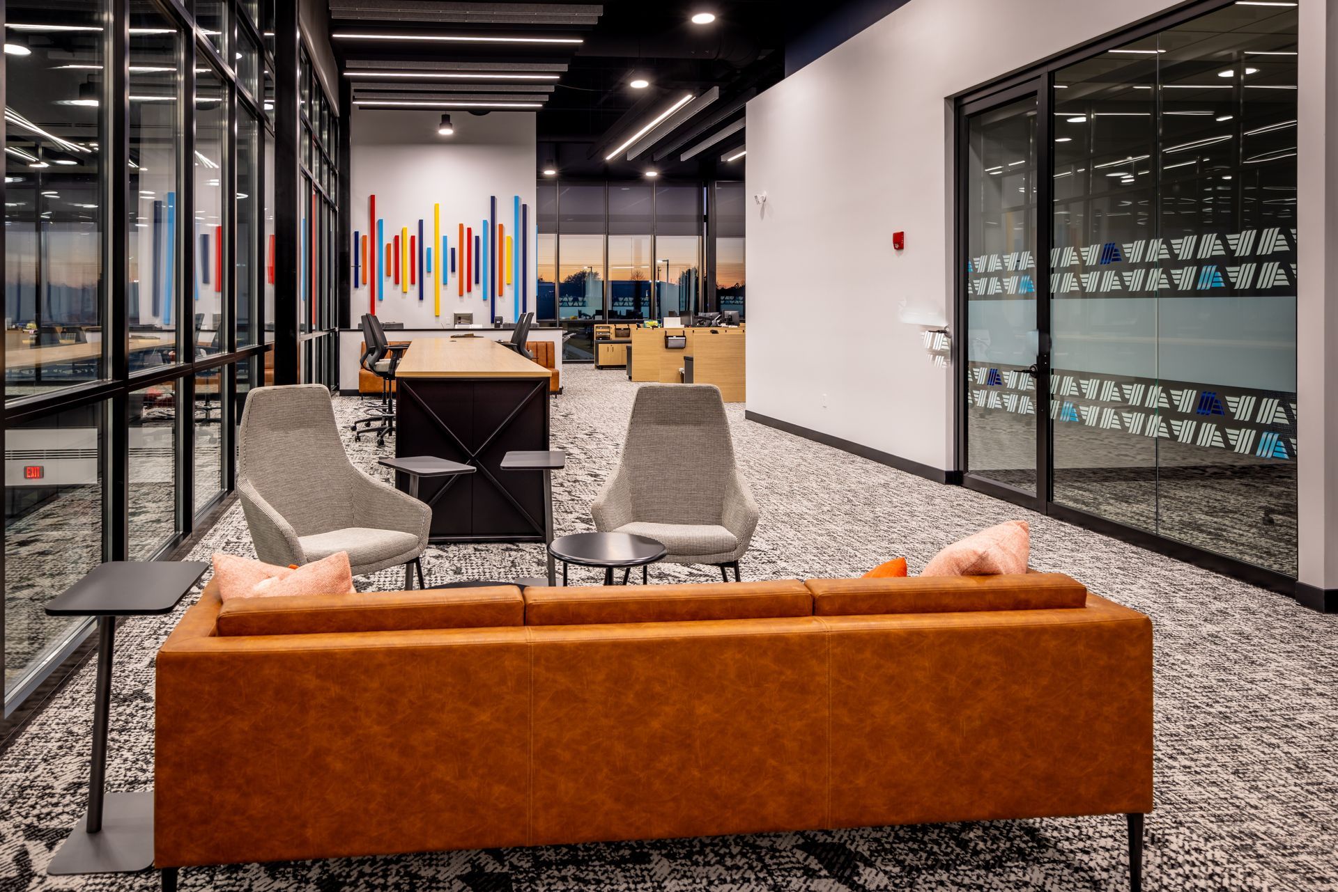 Modern office lounge with orange sofa, gray chairs, and glass-walled rooms around a central table.