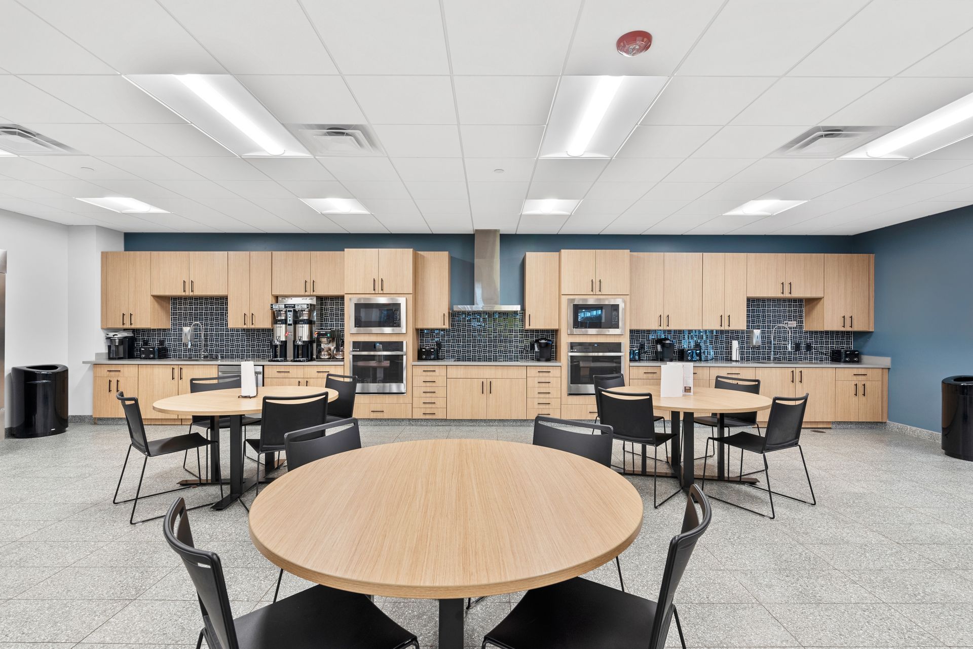 A modern break room with light wood cabinets, round tables, and black chairs. Blue accents and neutral flooring.
