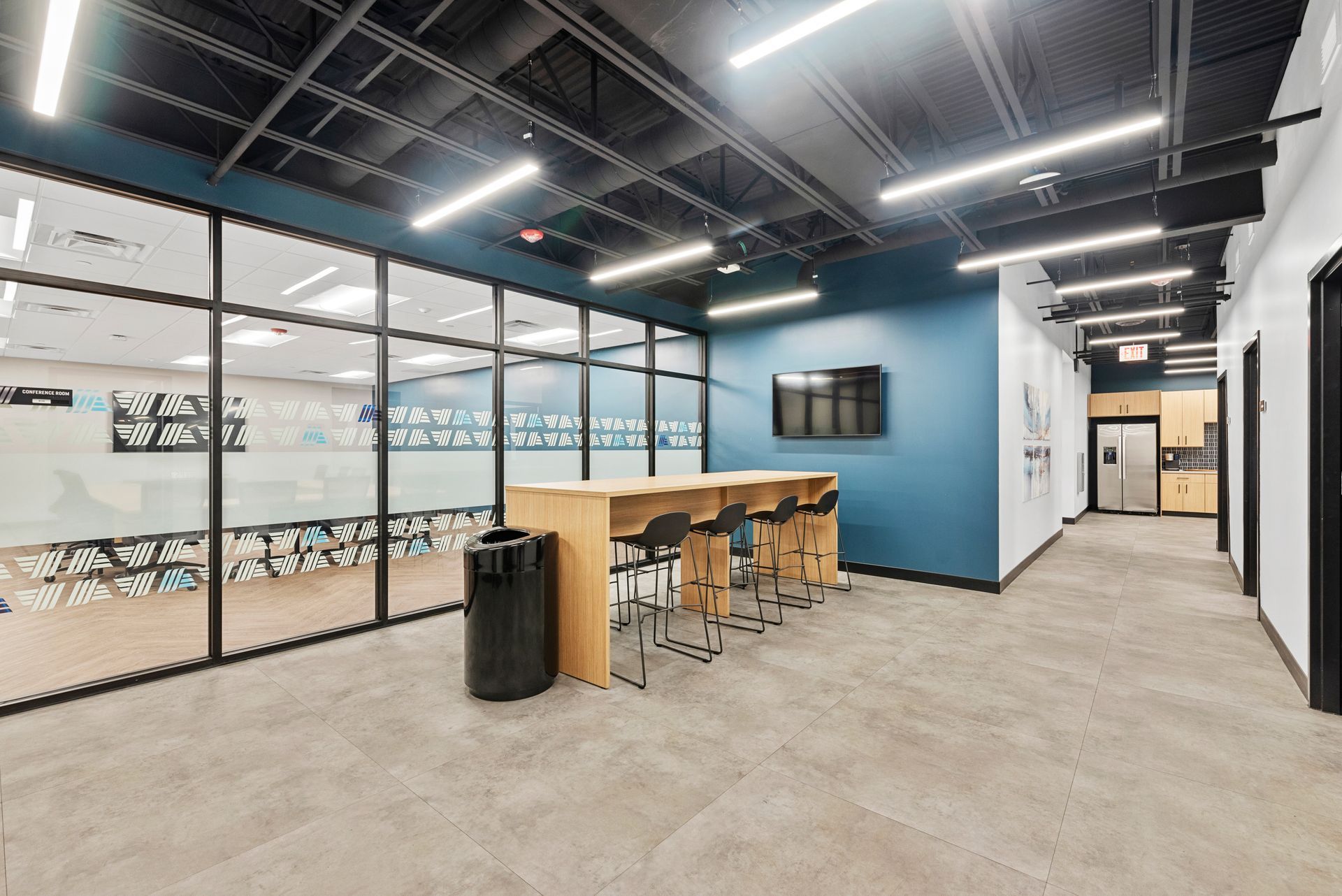Modern office break room with blue wall, glass-walled conference area, and high-top table with stools.