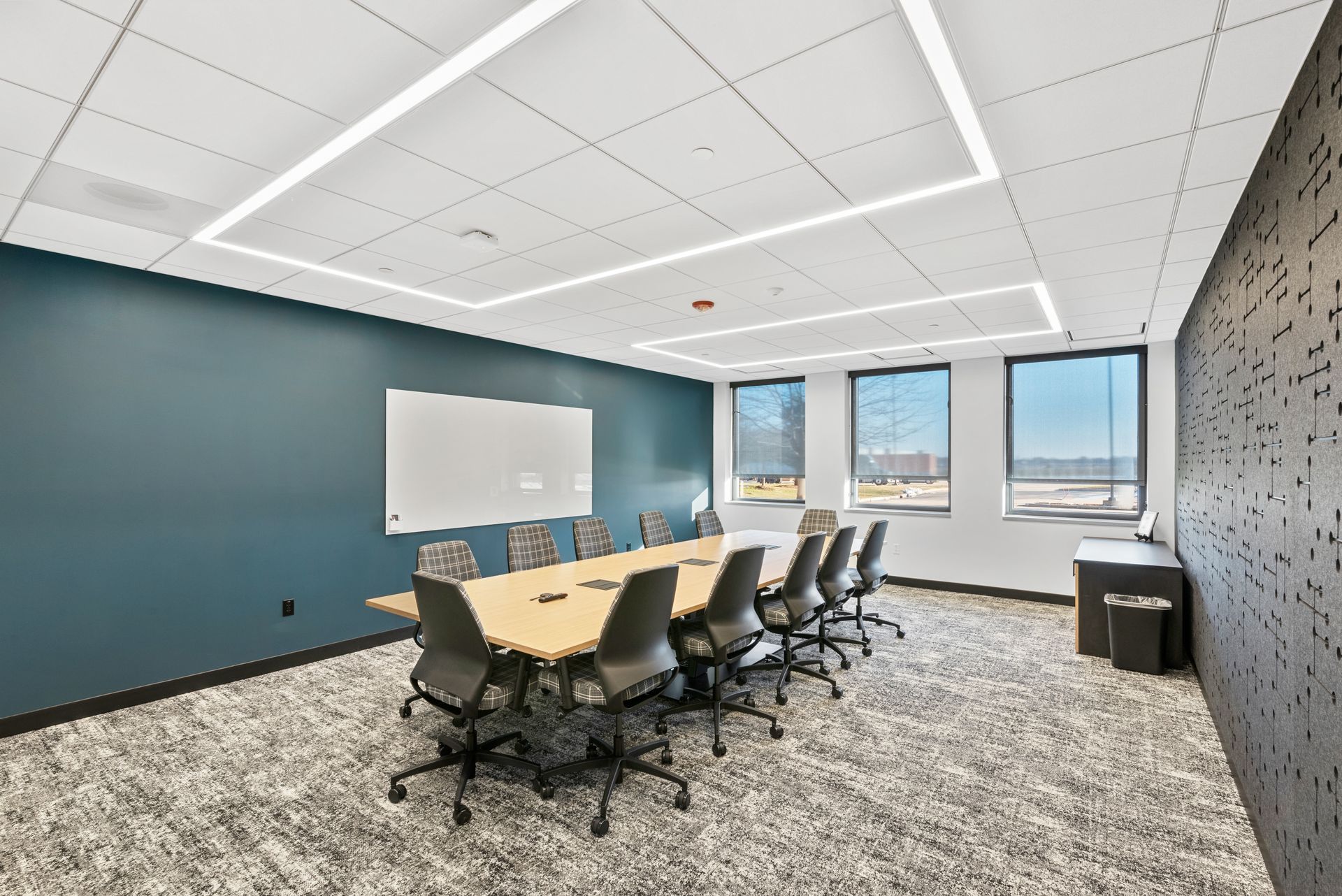 Conference room with a long table, chairs, whiteboard, windows, and patterned carpet. Teal wall and a textured accent wall.