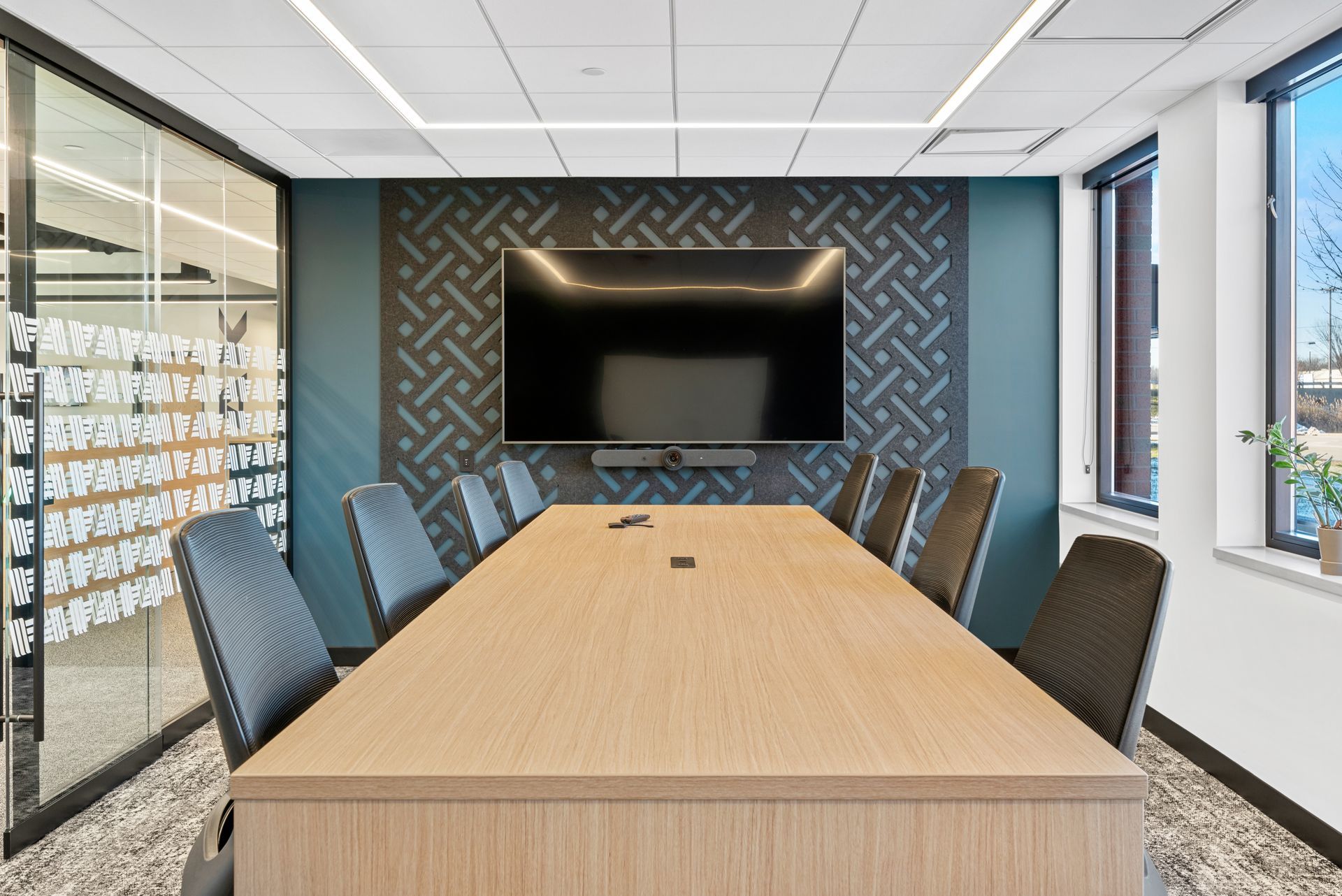 Conference room with large screen, table, and chairs. Blue patterned wall behind screen, glass wall on left.