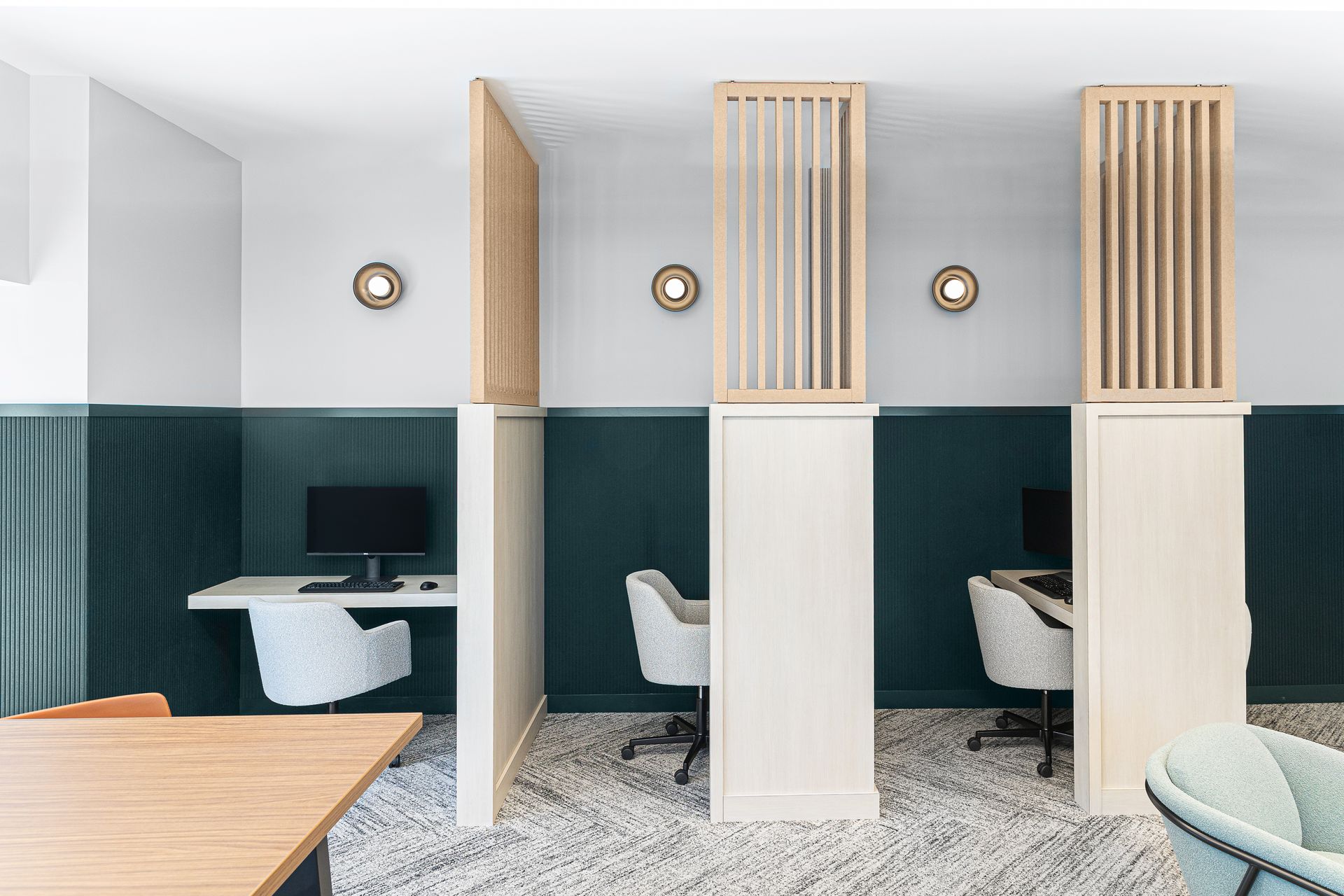 Office cubicles with desks and chairs, divided by wooden panels. White walls, dark green accent, and round wall lights.