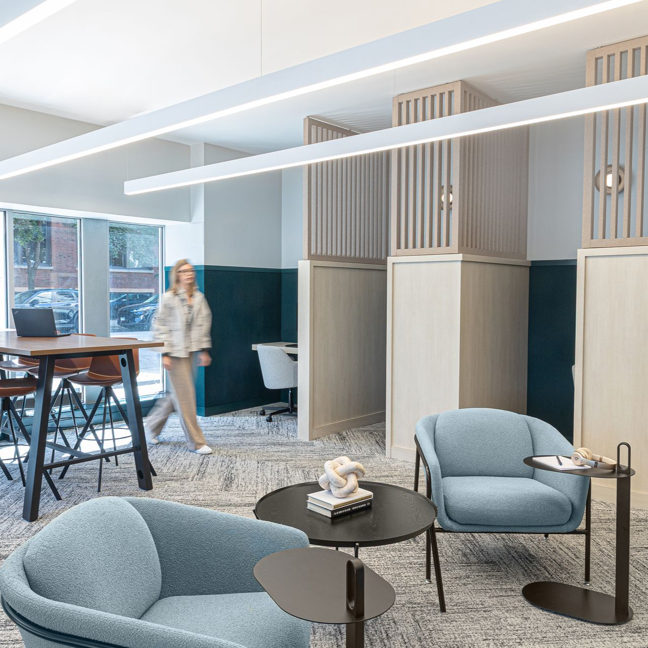 Co-working space: woman walks past blue armchairs, small tables, and a high-top table by a window.