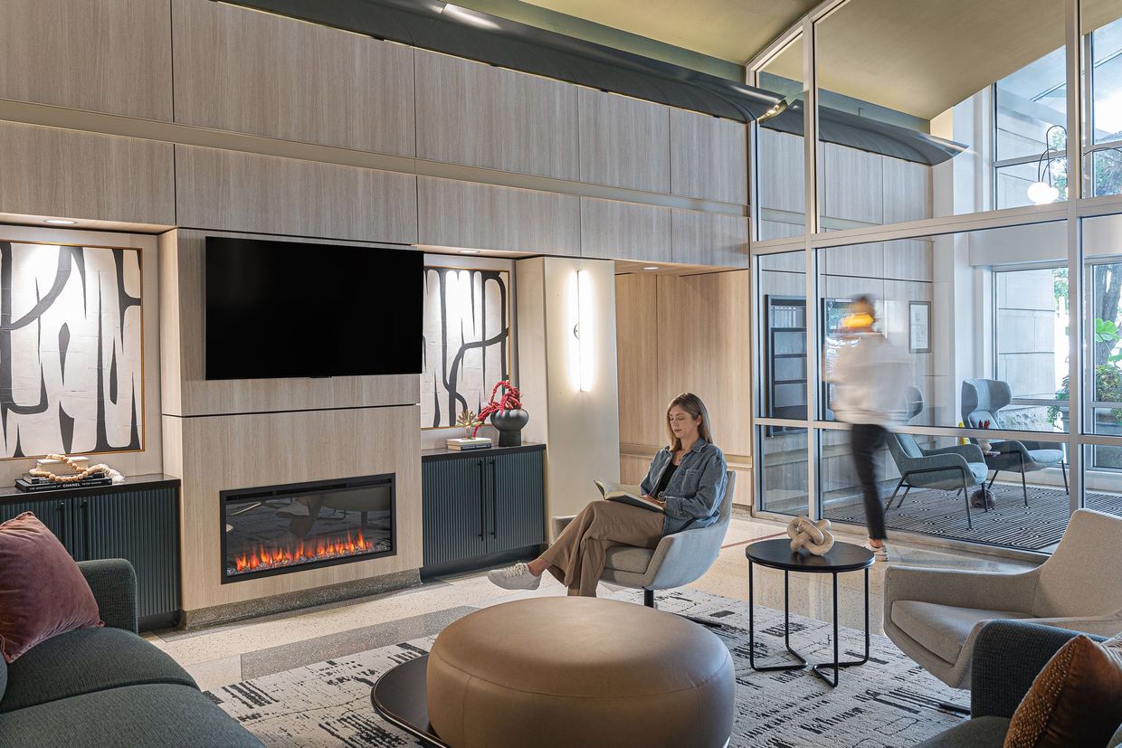 Lounge with fireplace, TV, and seated woman taking notes. Another person walks by glass wall.
