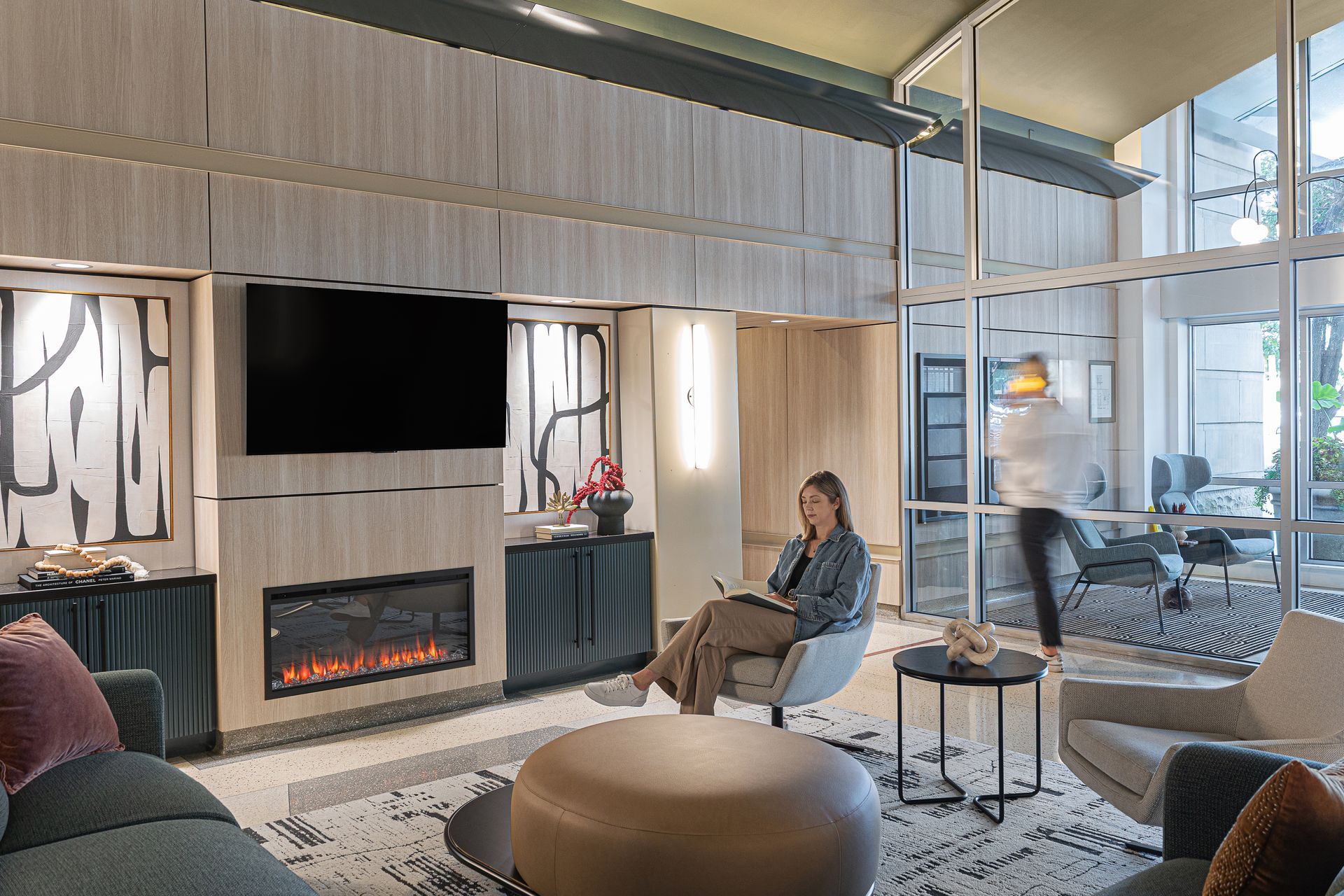 Lounge with fireplace, TV, and seated woman taking notes. Another person walks by glass wall.