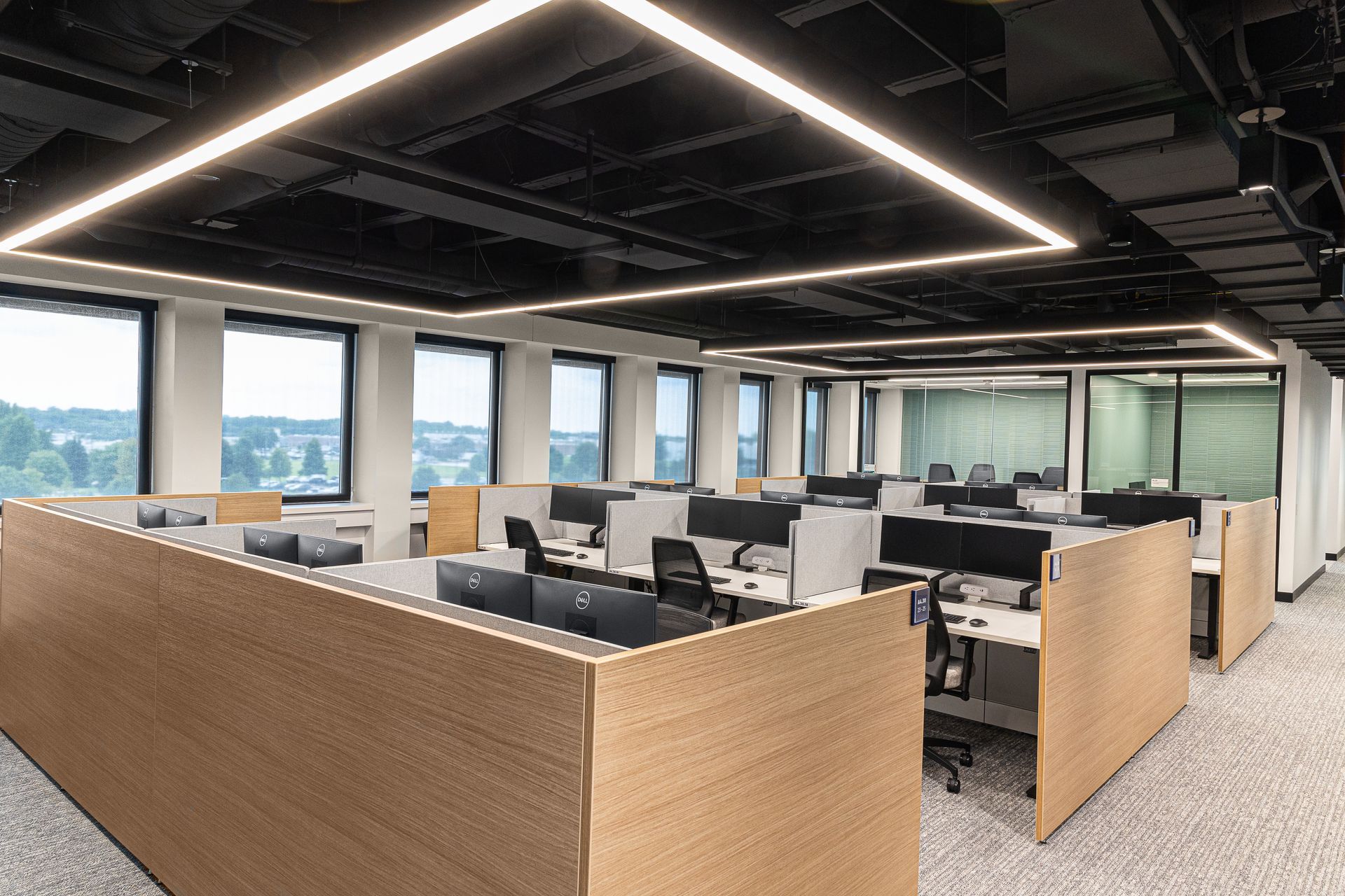 Open office with rows of desks, beige partitions, and large windows under a dark ceiling.