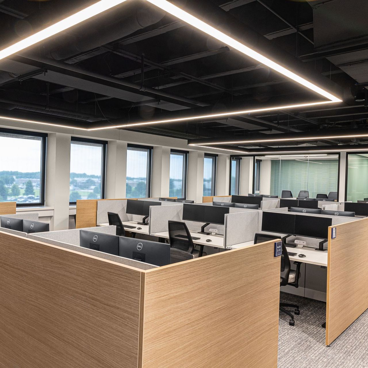 Modern open-plan office with cubicles, desks, large windows, and ceiling strip lights