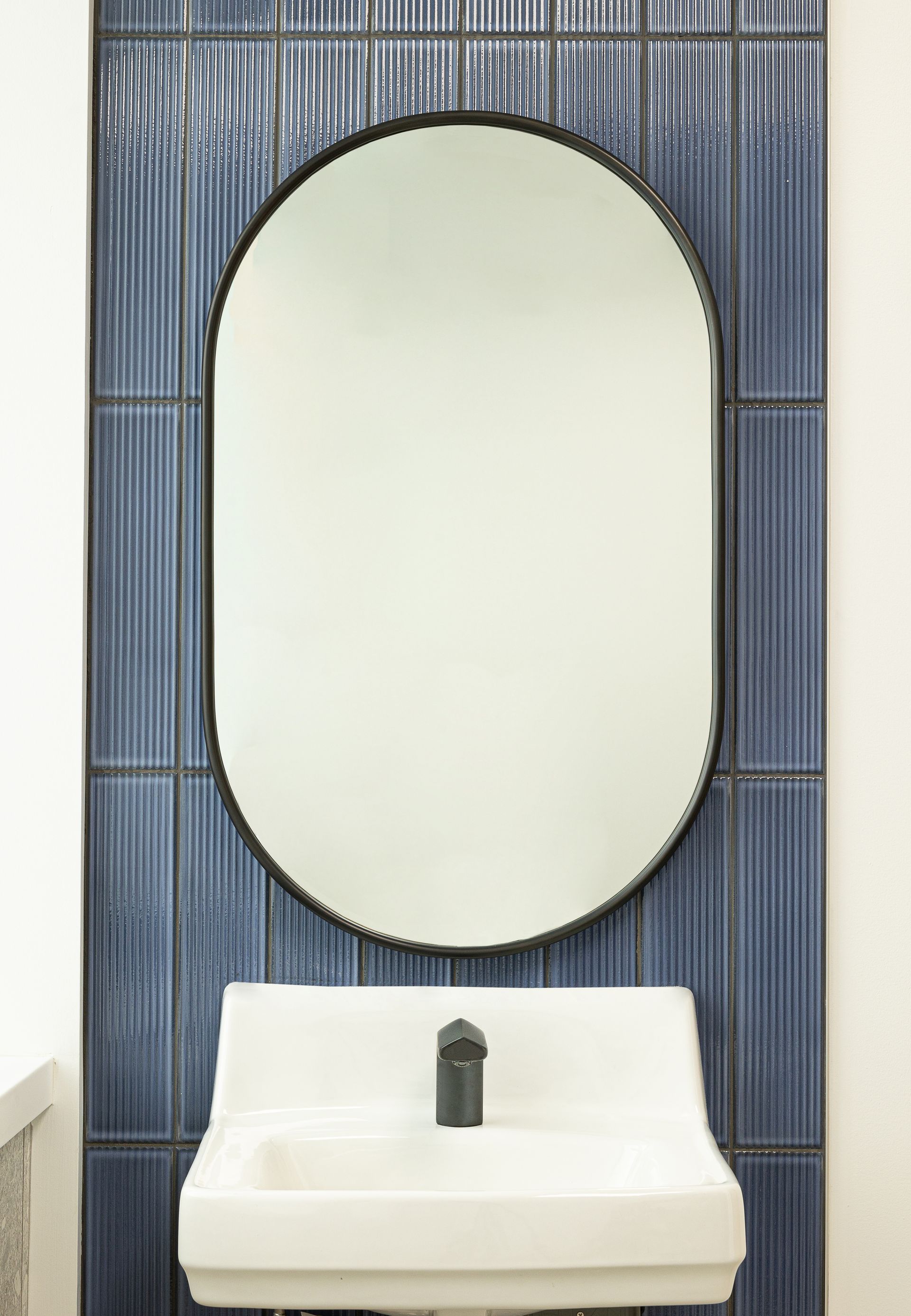Oval mirror over a white sink with a black faucet against a blue tile wall.