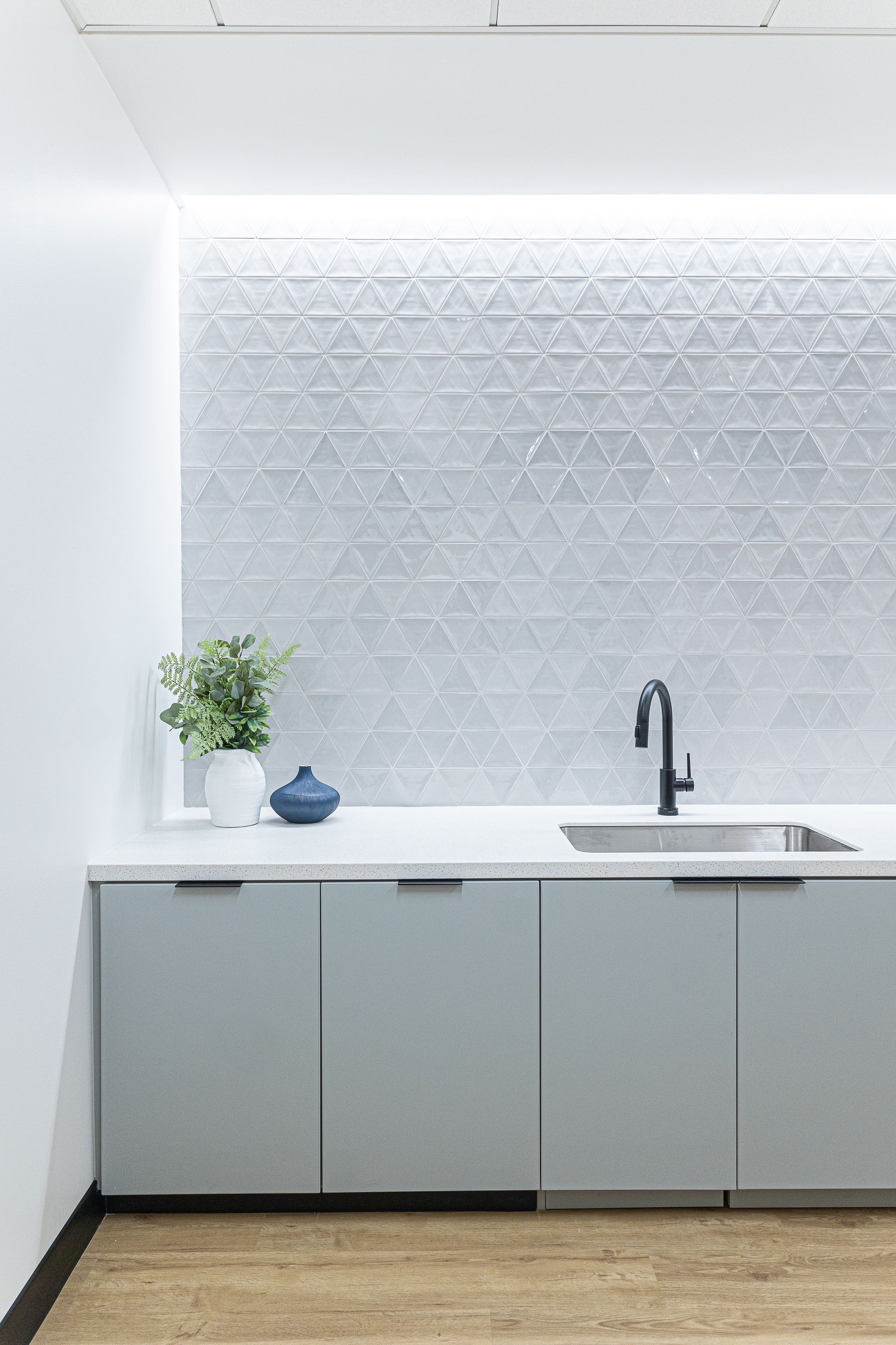 Light blue cabinets under a stainless steel sink with black faucet, triangle tile backsplash, and decorative plants.