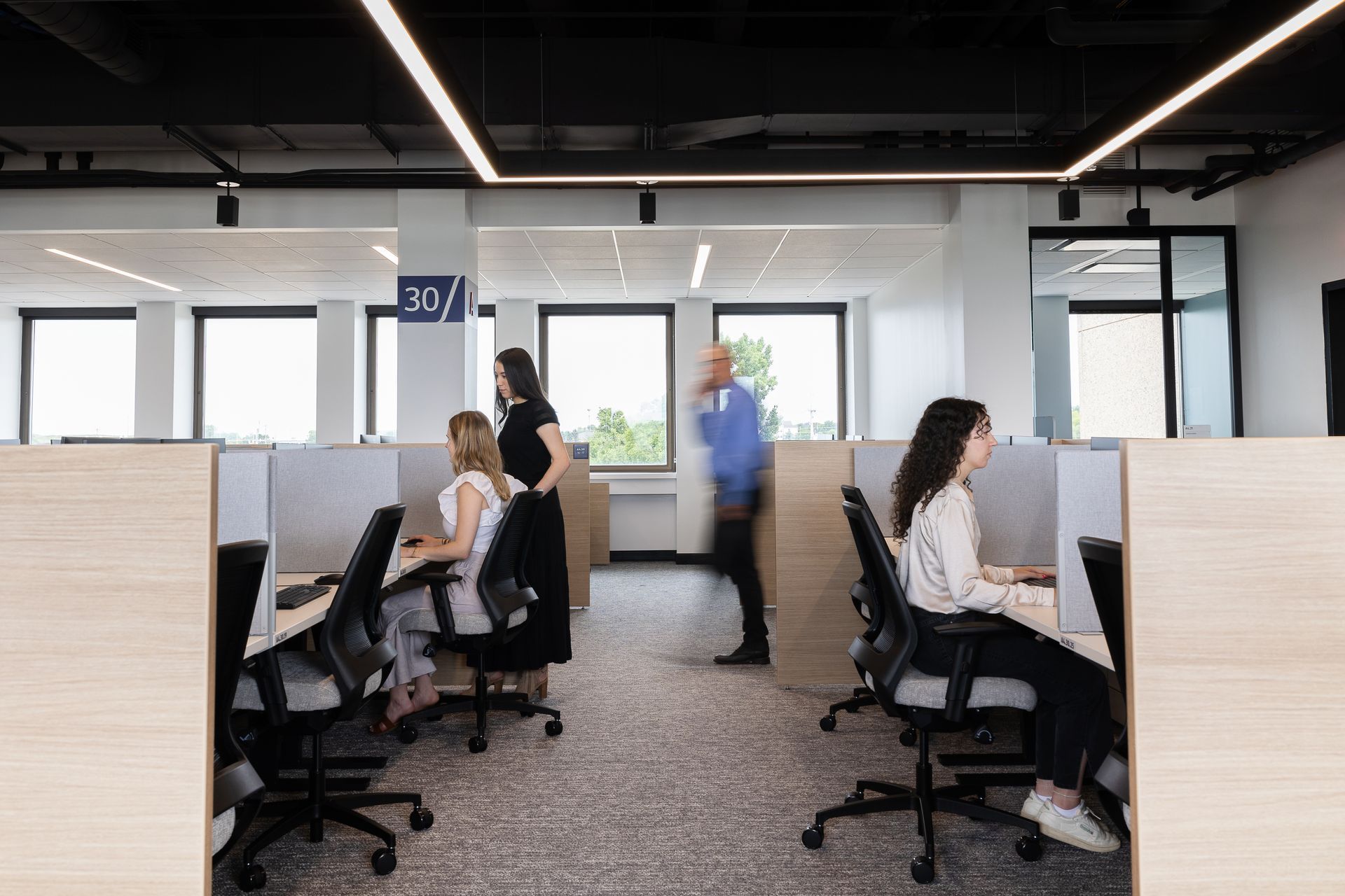 Office cubicles with people working; one person walks past, windows in background.