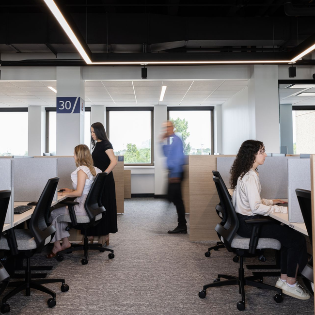Office space with people at desks, one talking on the phone, others working. Bright lighting.