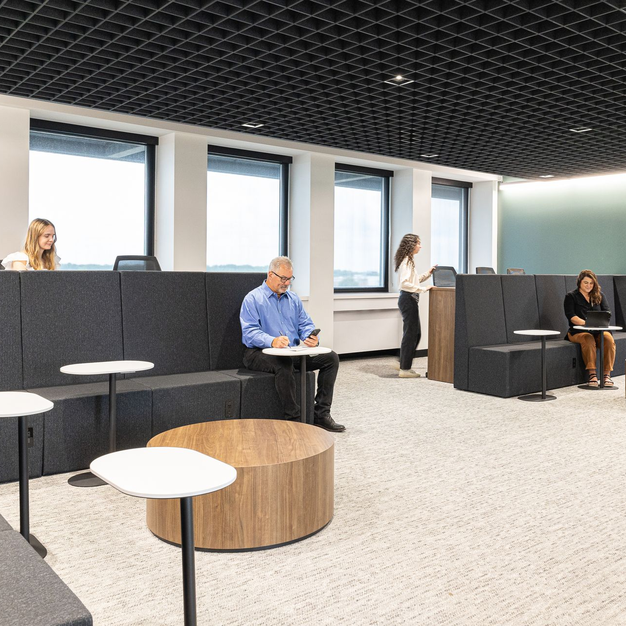 Office interior with people working at desks, casual seating, and large windows.
