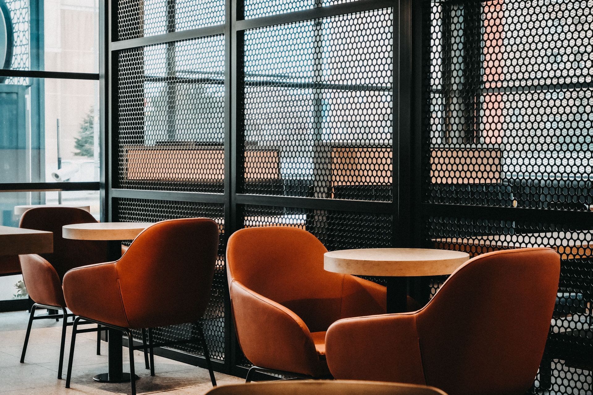 Modern café lounge with orange chairs, round tables, and a black mesh wall by large windows