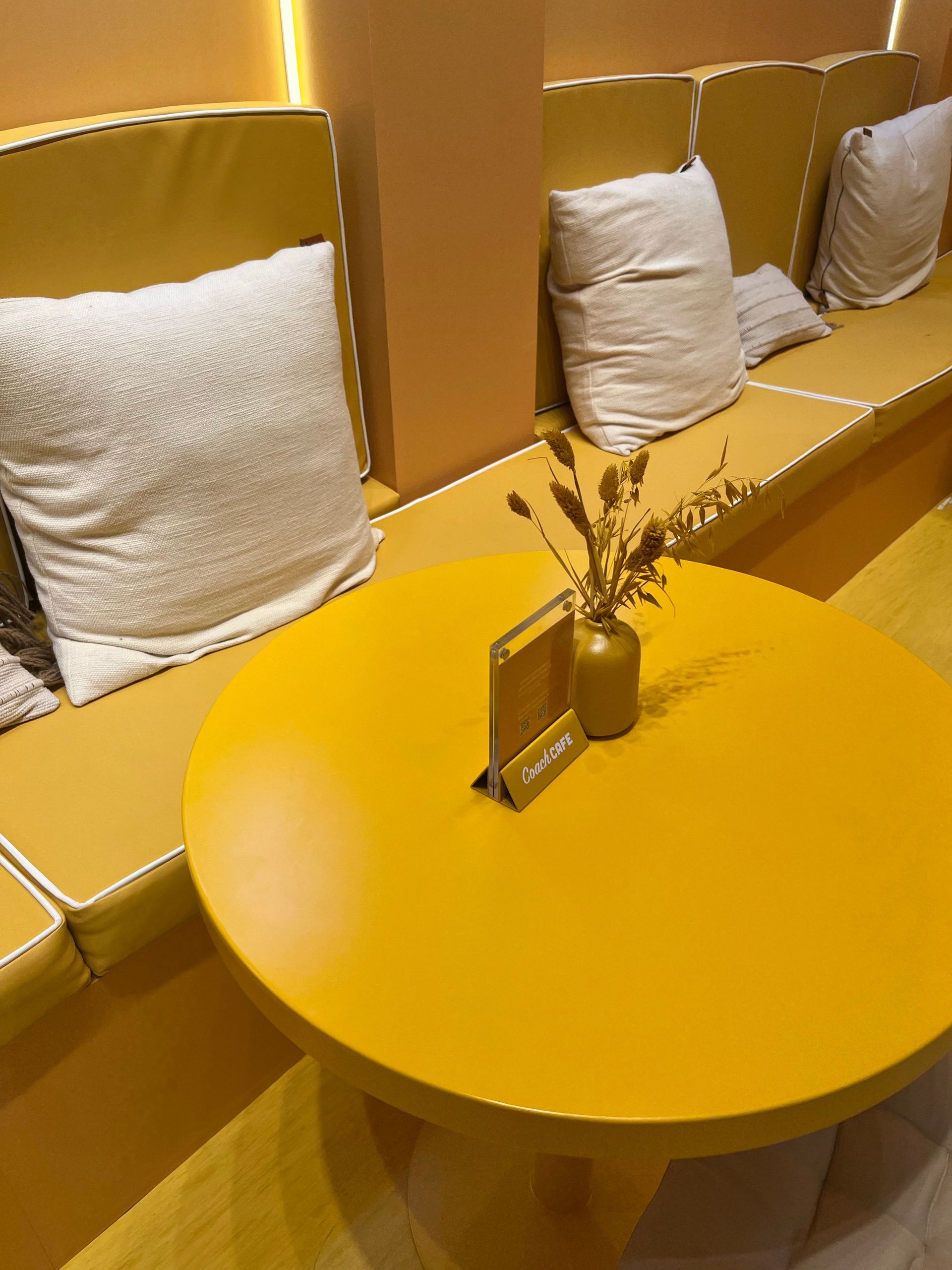 Yellow lounge with a round table, white pillows on a bench, and a small vase centerpiece.