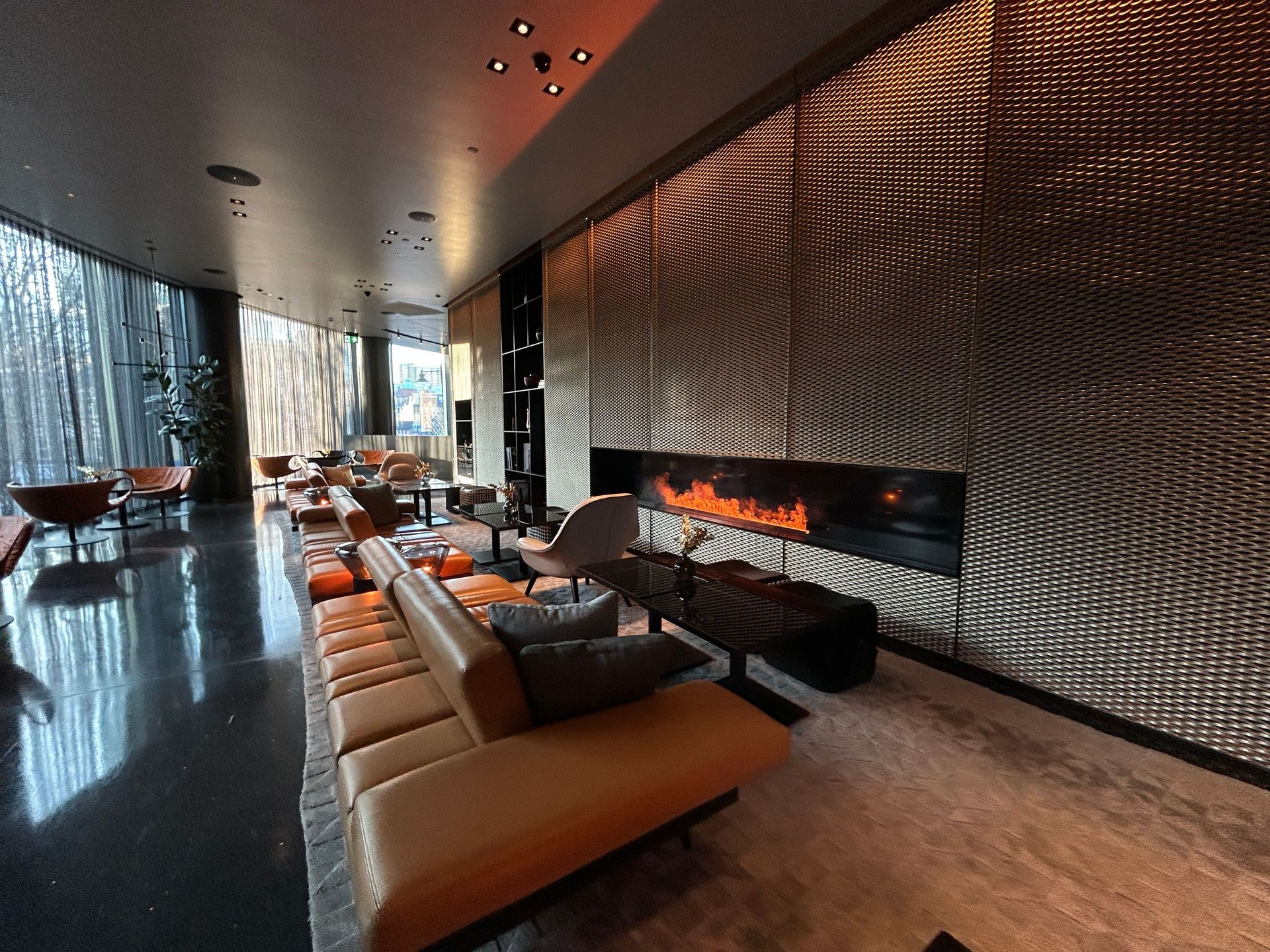 Modern lounge with tan sectional seating, metallic feature wall, and long fireplace by large windows