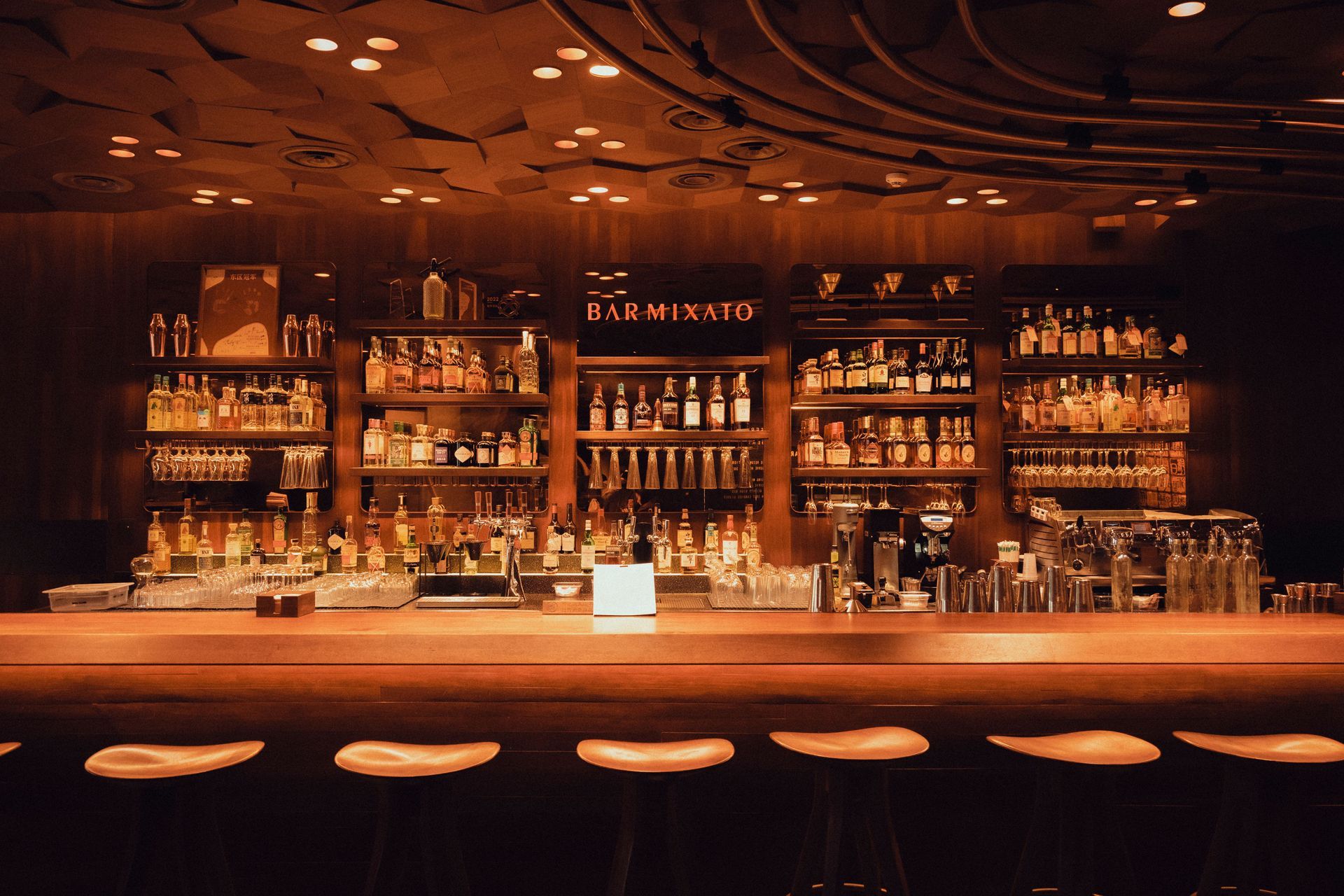 Dimly lit wooden bar interior with shelves of bottles and a row of stools at the counter