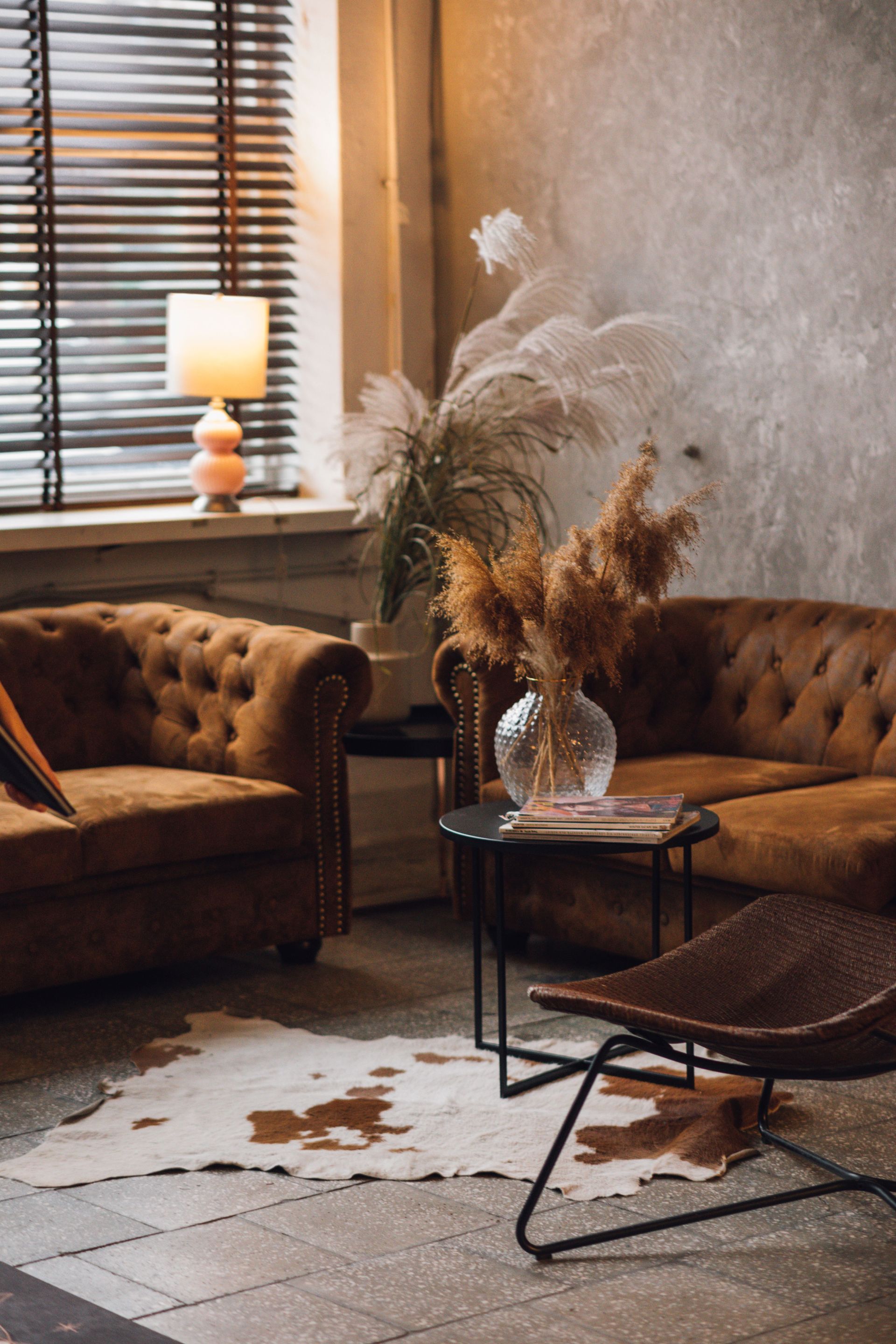 Cozy living room with brown sofas, round table, dried flowers, lamp, and window blinds