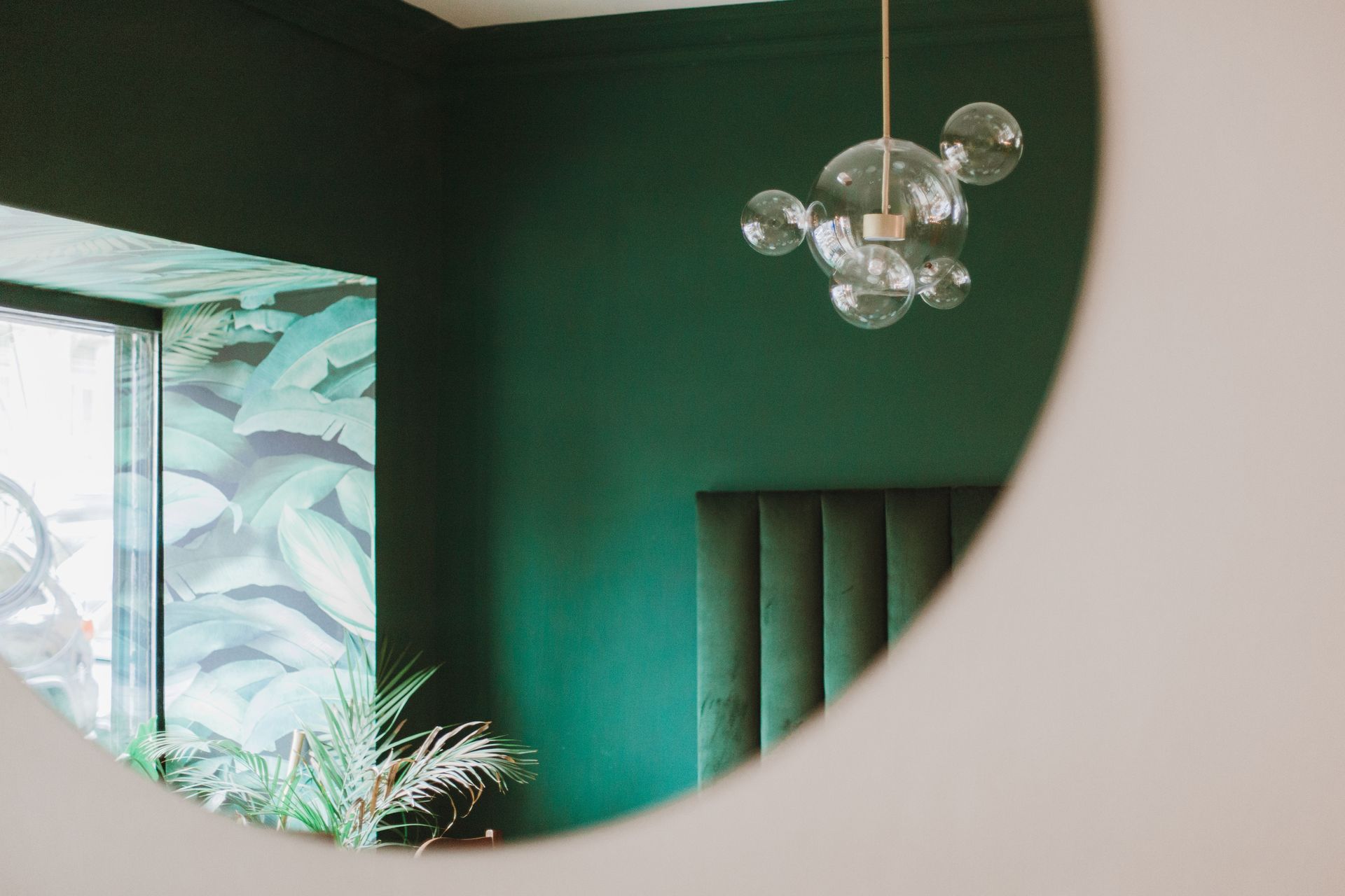 Round mirror reflecting a green-walled room with a silver globe chandelier and a dark tufted chair