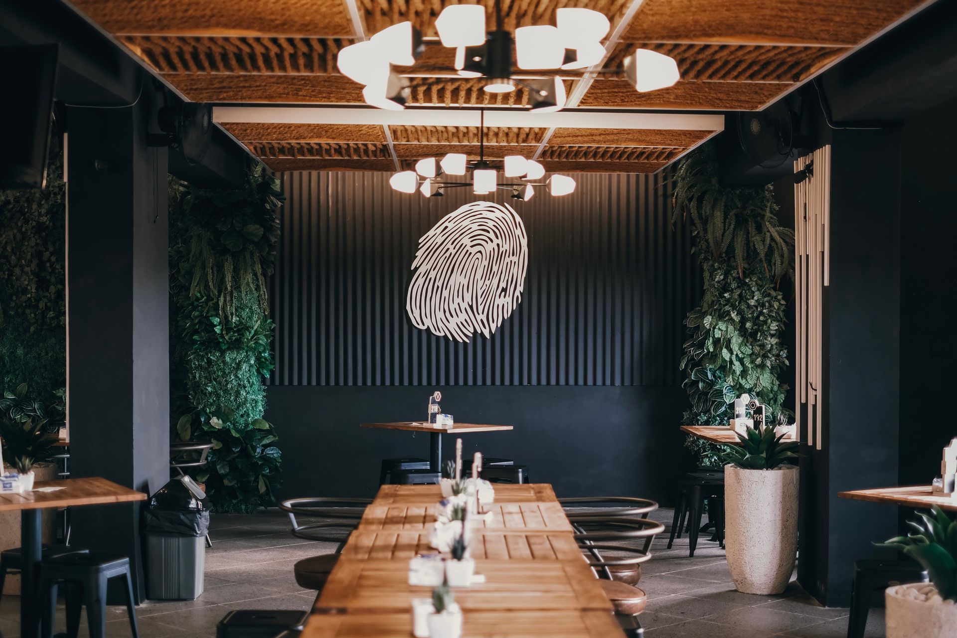 Modern restaurant dining room with a long wooden table, black walls, hanging lights, and potted greenery