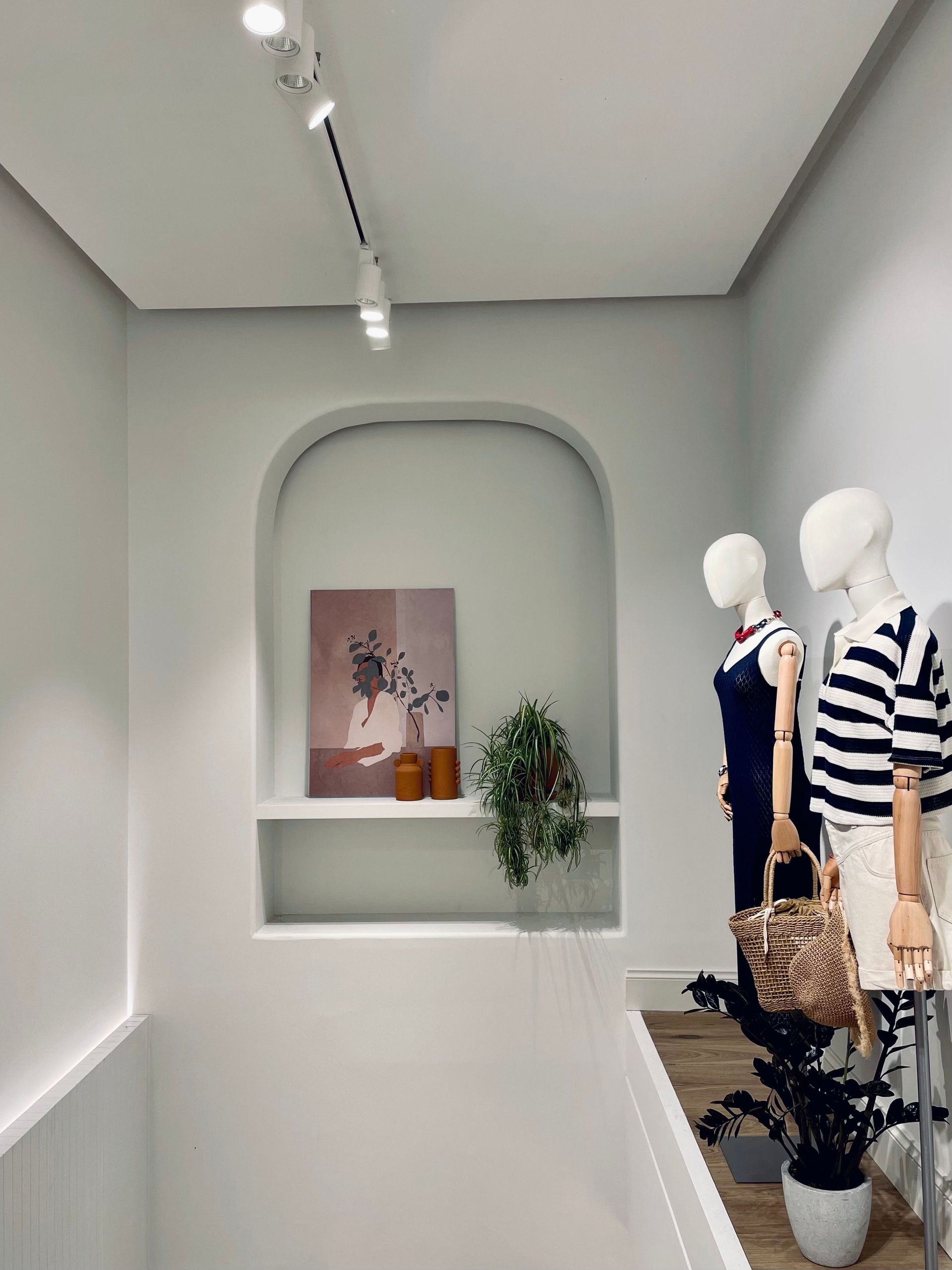 Minimalist clothing store display with mannequins, arched niche, and potted plant in a gray-white room