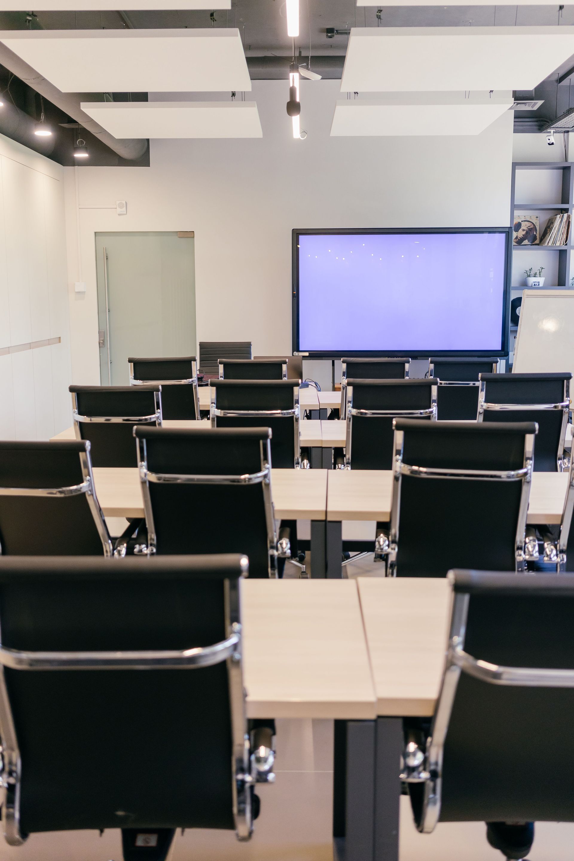 Empty classroom with rows of black chairs and desks facing a large projector screen