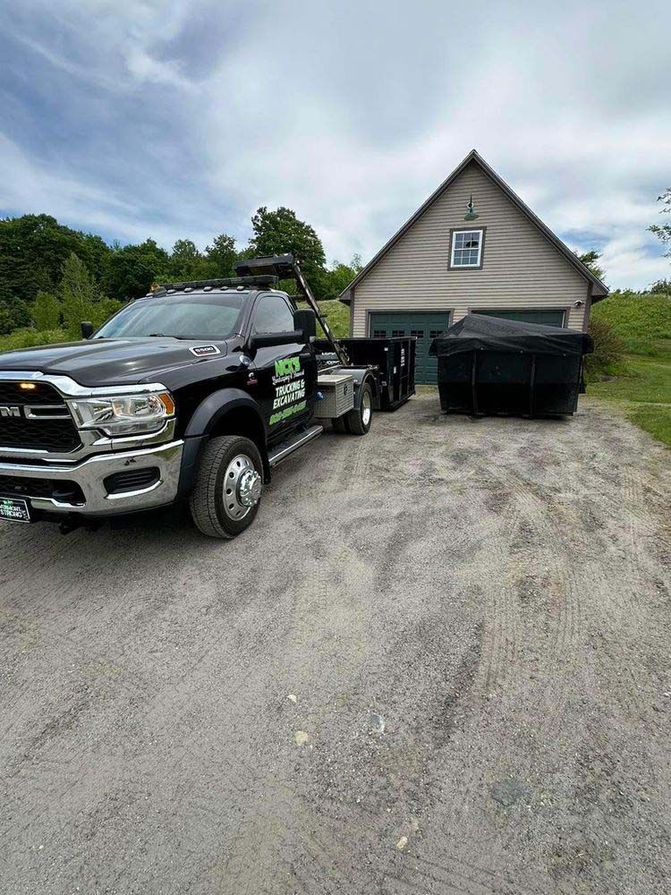 A black truck is towing a dumpster in front of a garage.