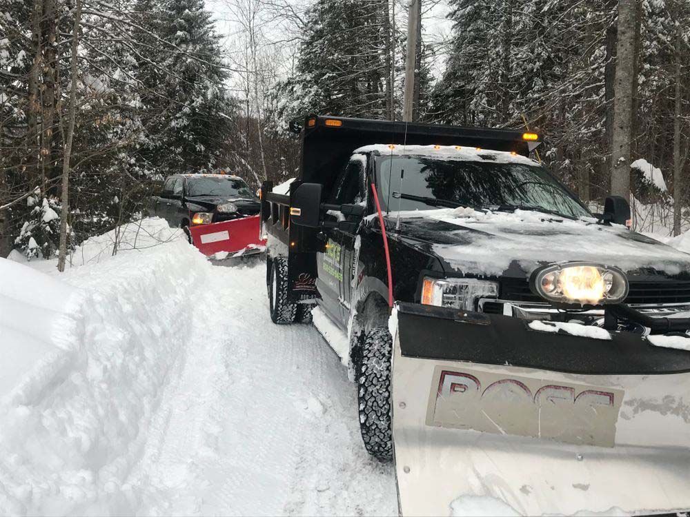 A snow plow is driving down a snowy road next to a dump truck.