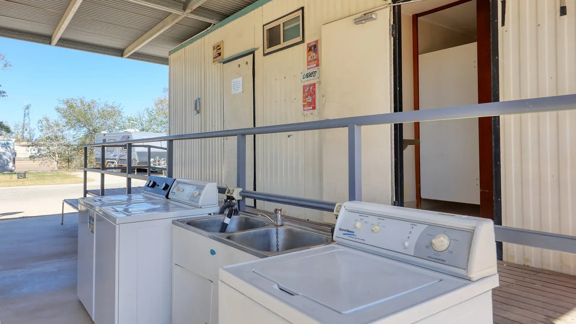 Outdoor laundromat with three white washing machines under a covered awning.