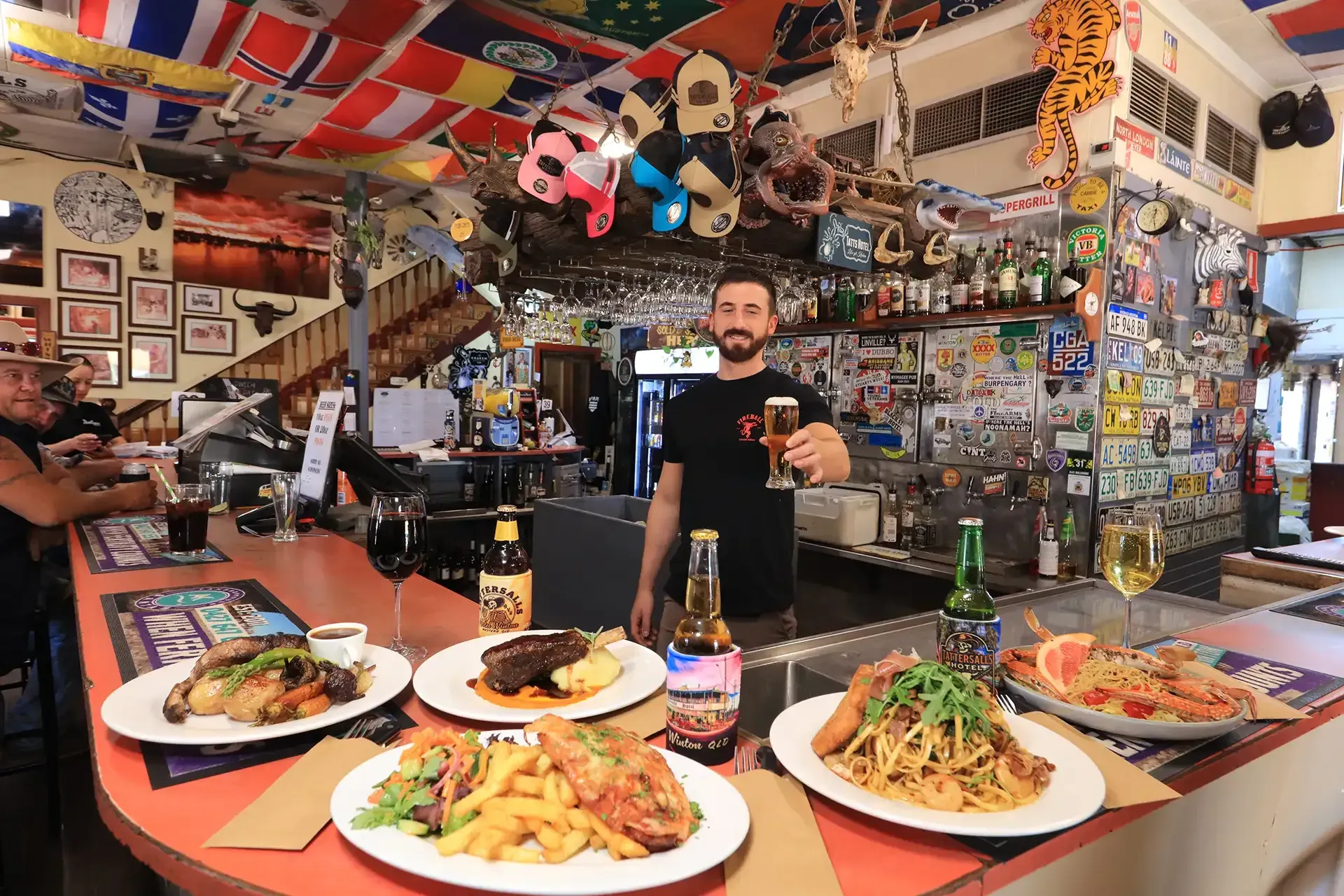 Man behind a bar counter with plates of food and drinks in a colorful, flag-filled restaurant