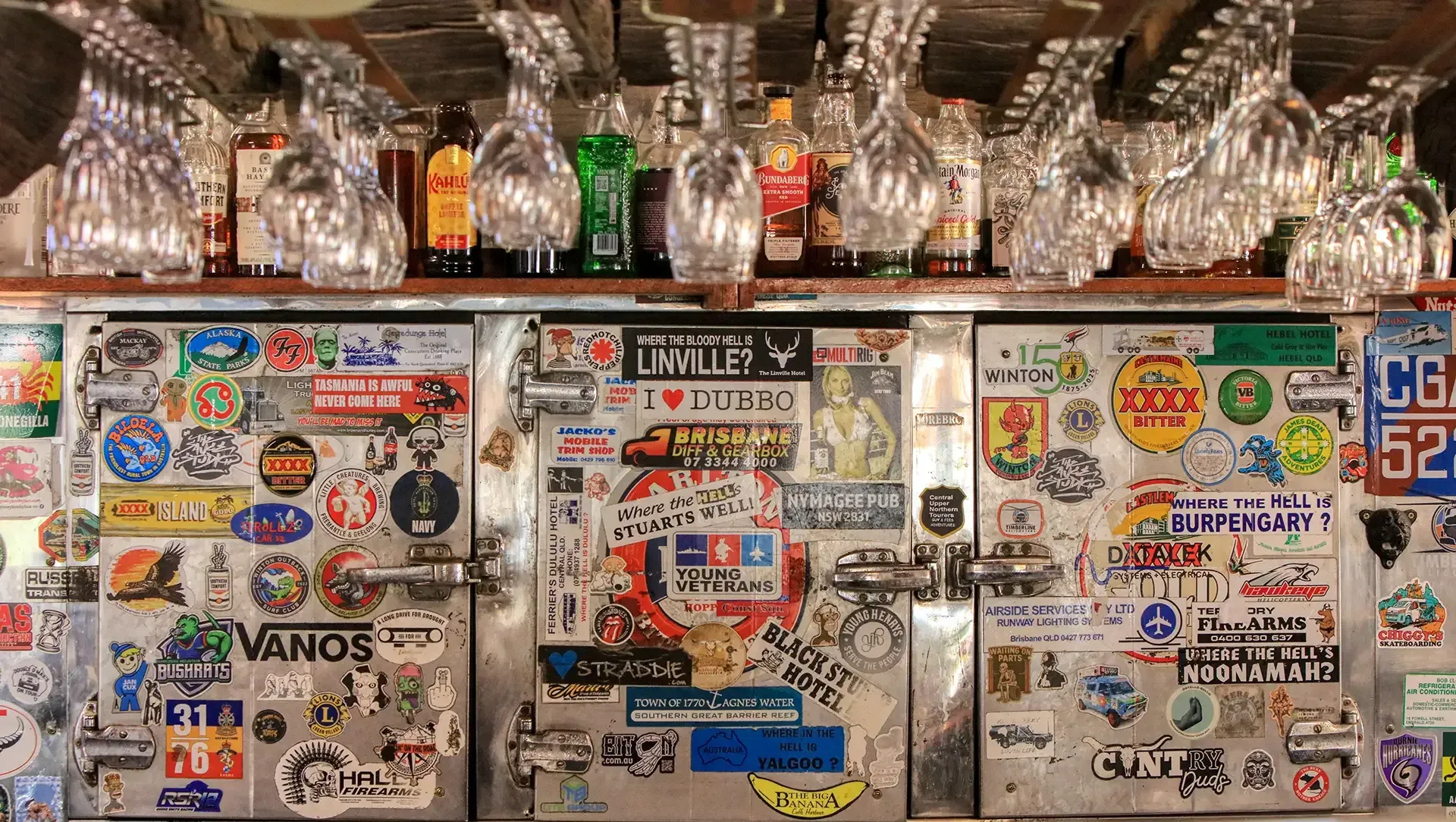 Sticker-covered bar shelf with hanging glasses and liquor bottles above a graffiti-covered counter