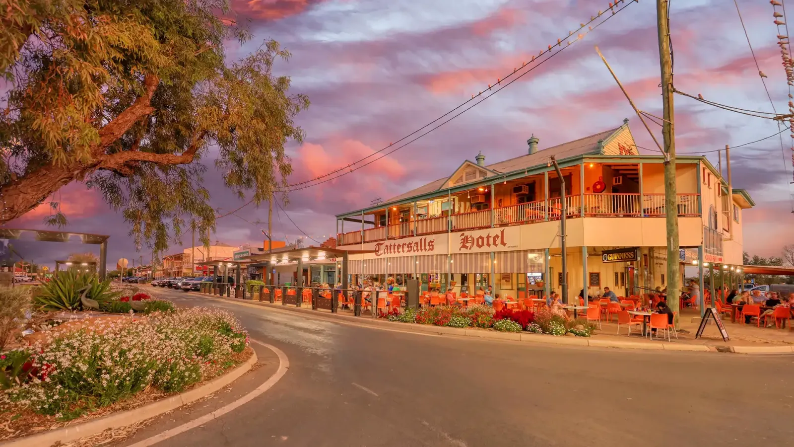 Curving street beside a pink two-story café with outdoor seating, string lights, and flowers at sunset.
