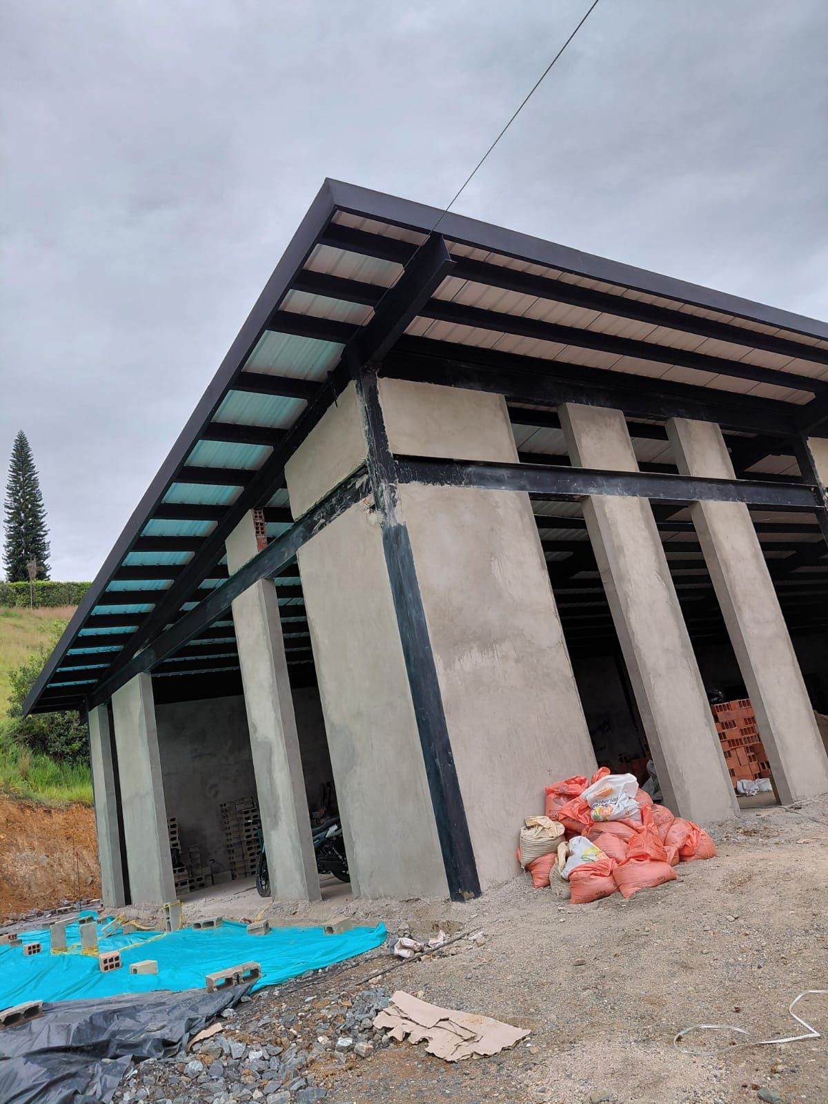 Estructura de hormigón parcialmente construida con vigas de techo oscuras sobre un terreno de grava, lona azul y escombros en las cercanías.