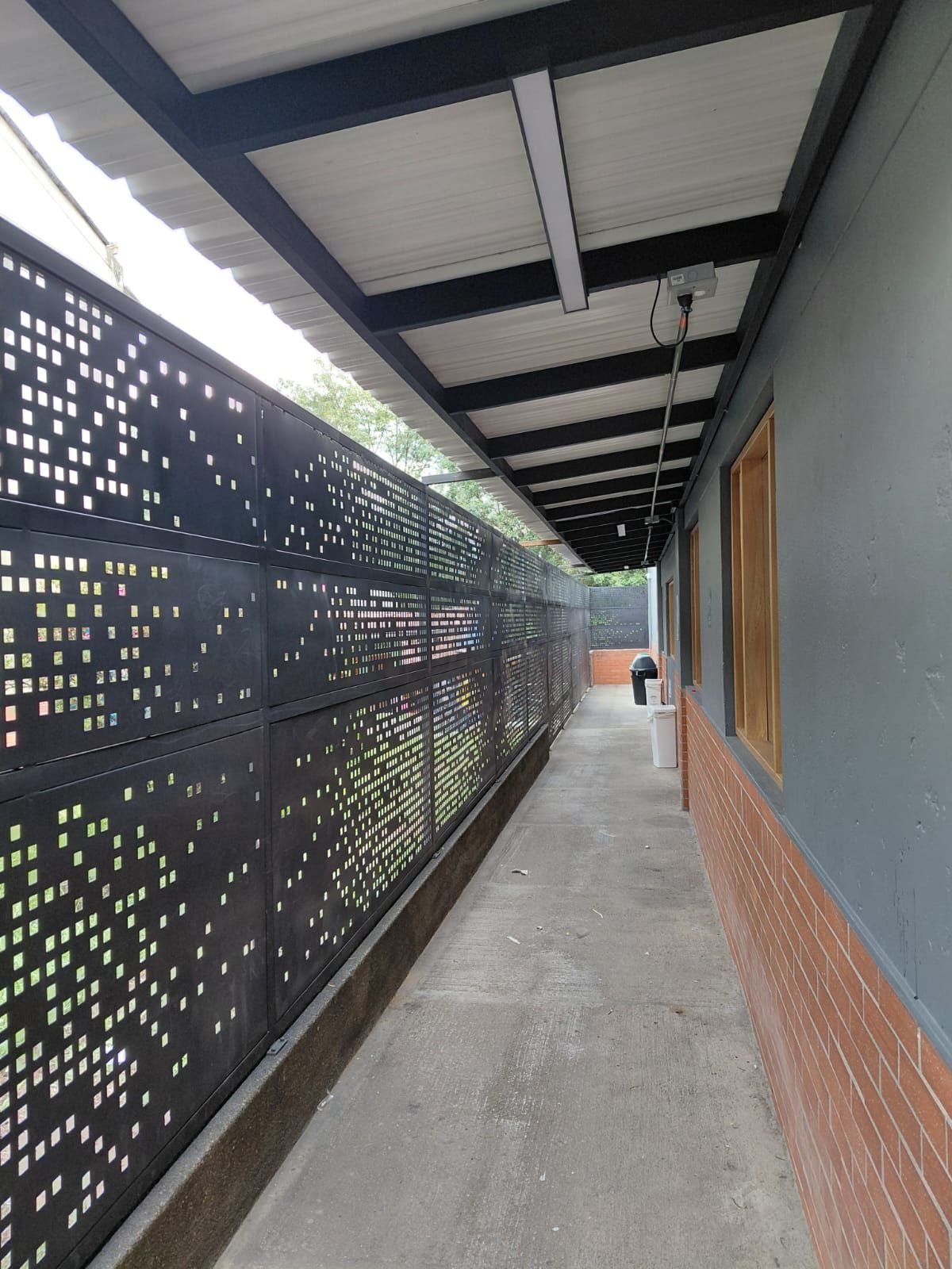 Corredor exterior cubierto con valla negra perforada, pared gris y suelo de ladrillo que se extiende hacia el horizonte.