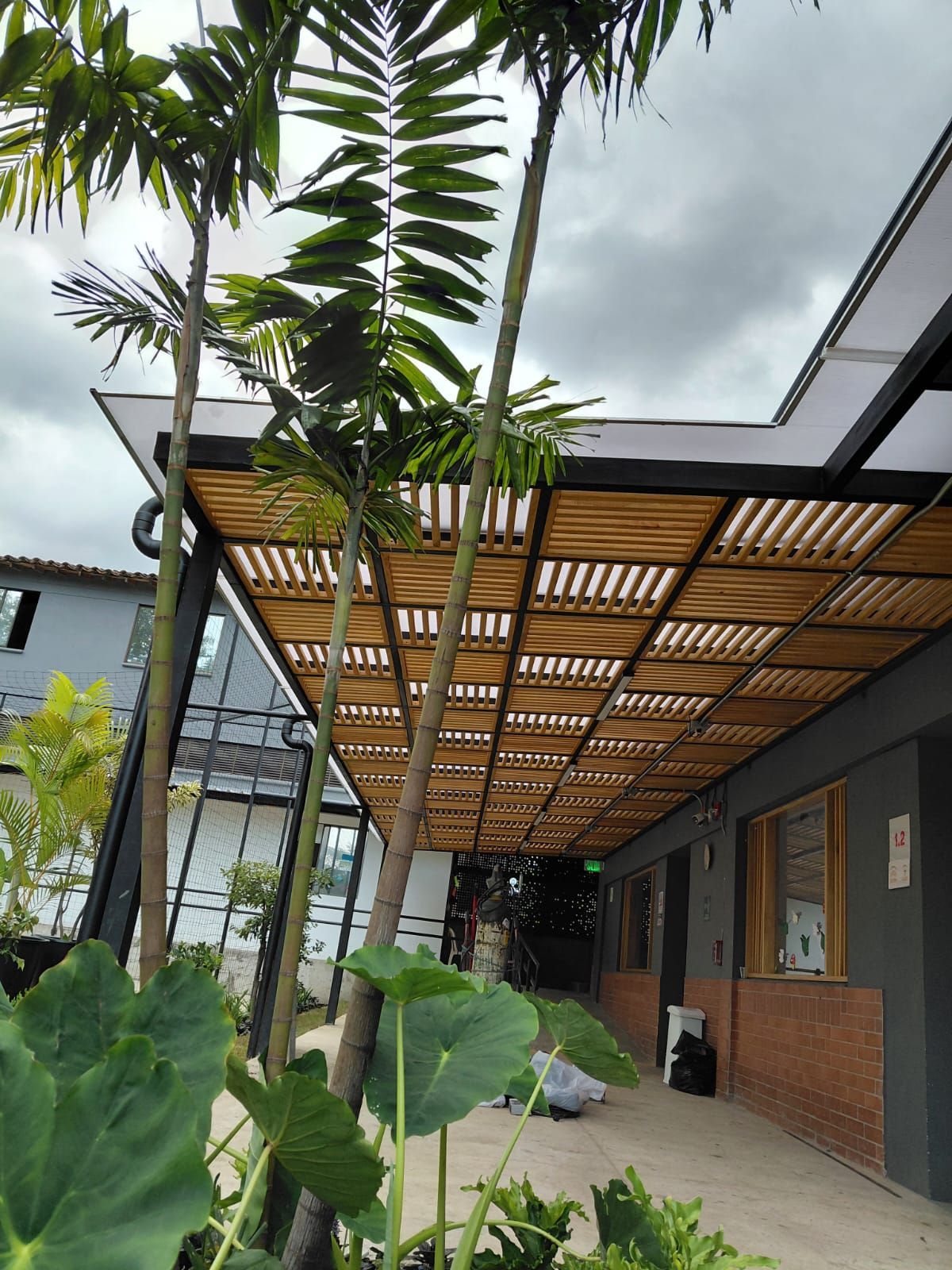 Pasarela cubierta con techo de listones de madera, paredes de ladrillo y exuberantes plantas tropicales bajo un cielo nublado.