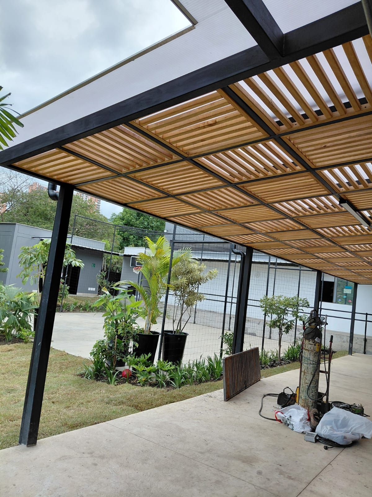 Pasarela exterior cubierta con postes negros, techo de listones de madera y materiales de construcción sobre un suelo de hormigón.