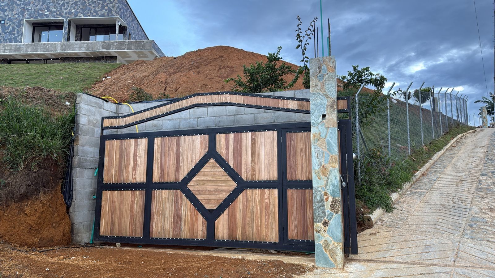 Puerta de madera con diseño de diamante junto a un camino de tierra y una valla metálica bajo un cielo nublado.