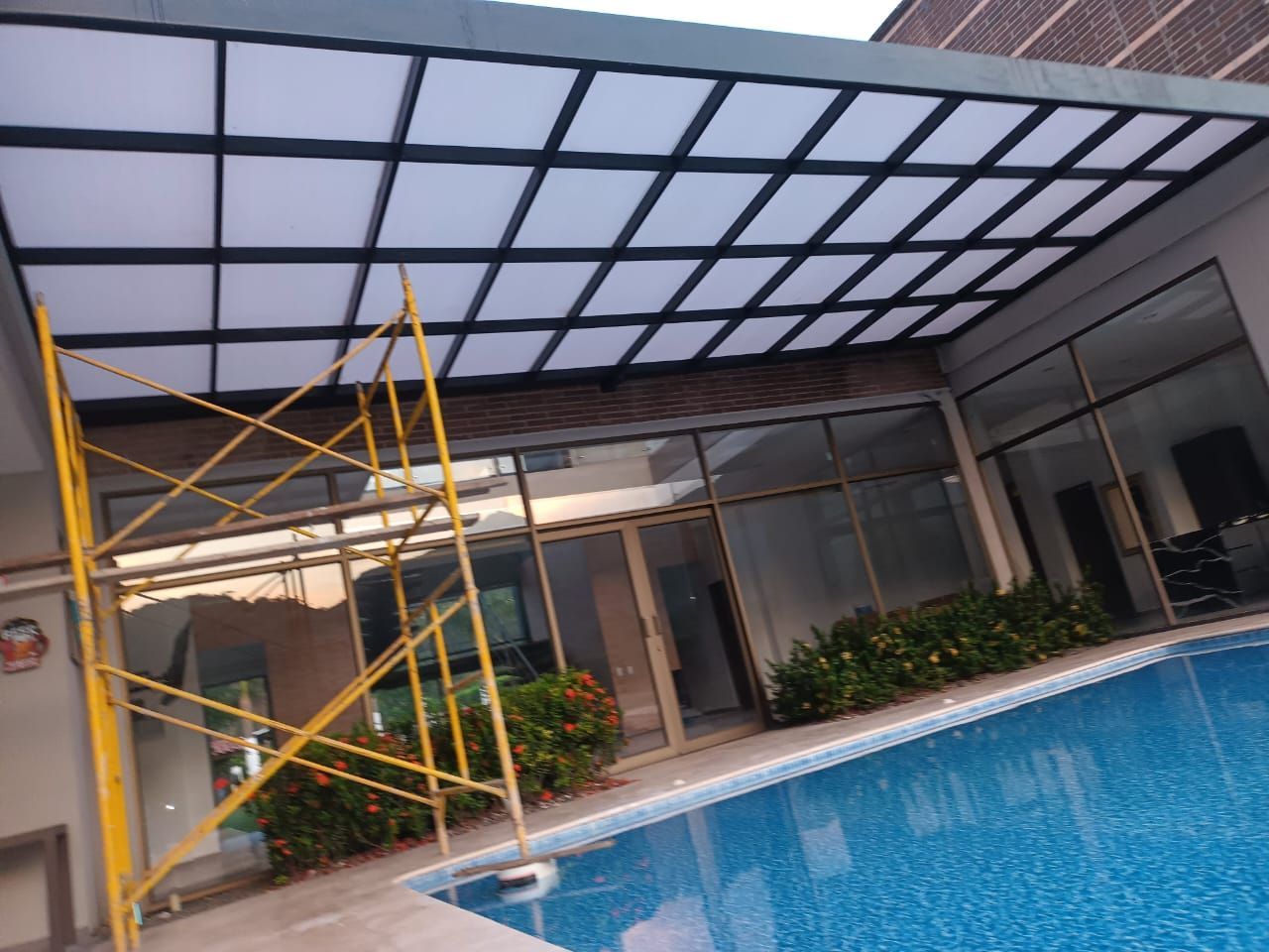 Piscina cubierta interior bajo un techo de cristal, con andamios amarillos junto al agua y puertas de cristal en la parte trasera.