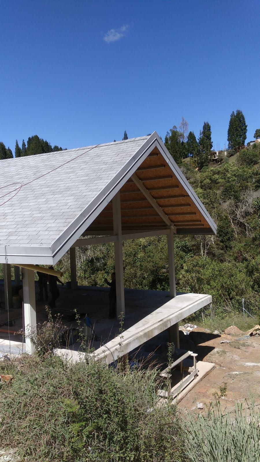 Pequeño refugio abierto por los lados, con techo inclinado y banco, situado en una ladera bajo un cielo azul despejado.