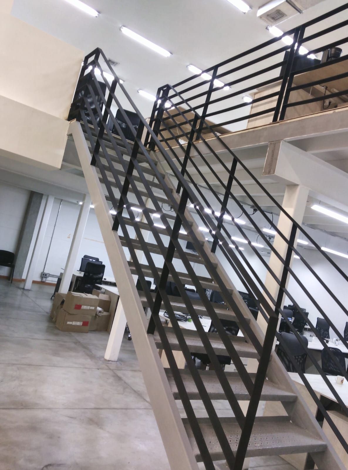 Escalera de metal negro en un luminoso interior de oficina con barandillas de cristal y cajas cerca.