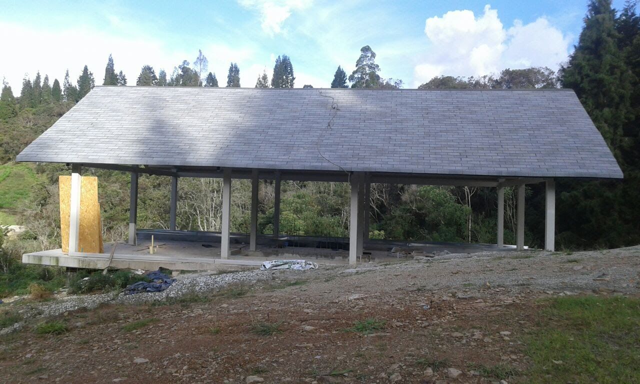 Pabellón al aire libre con techo gris y pilares de hormigón en una zona rural boscosa.