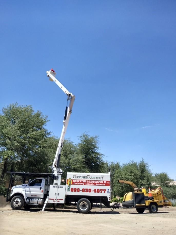 White bucket truck with boom lift raised beside a yellow woodchipper on a dirt lot under blue sky