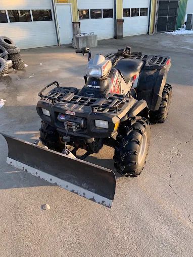 Black ATV with a front plow parked on a concrete lot outside a building.